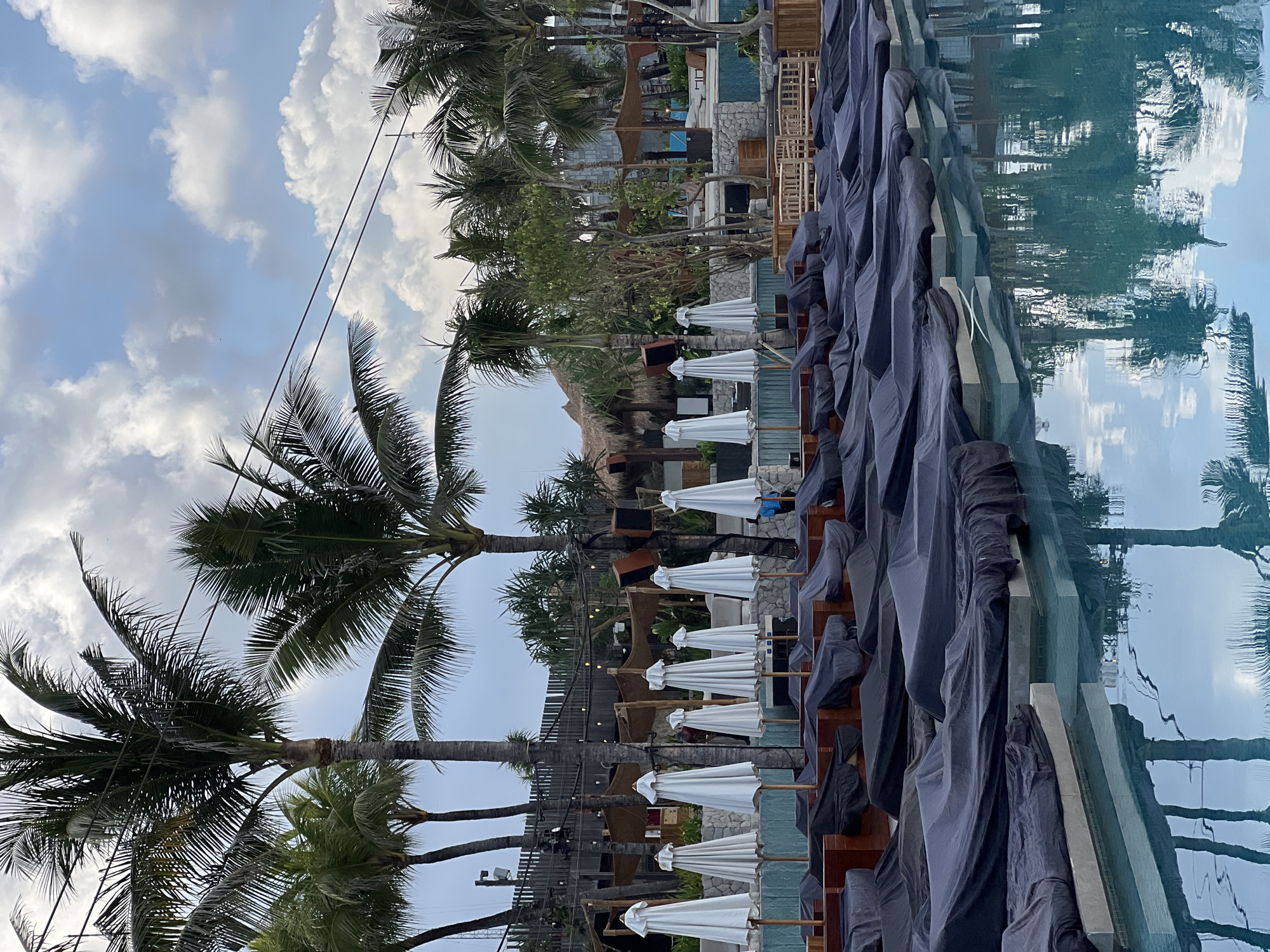 The image depicts an outdoor swimming pool area at a resort in North Kuta, Indonesia. Numerous lounge chairs are covered in dark grey tarps, suggesting they are not in use. White closed umbrellas stand in rows behind the lounge chairs. Palm trees are abundant in the background, and the sky is partly cloudy. A person in a blue shirt is visible in the background near some stone structures. The water of the pool reflects the surrounding palm trees and the sky.