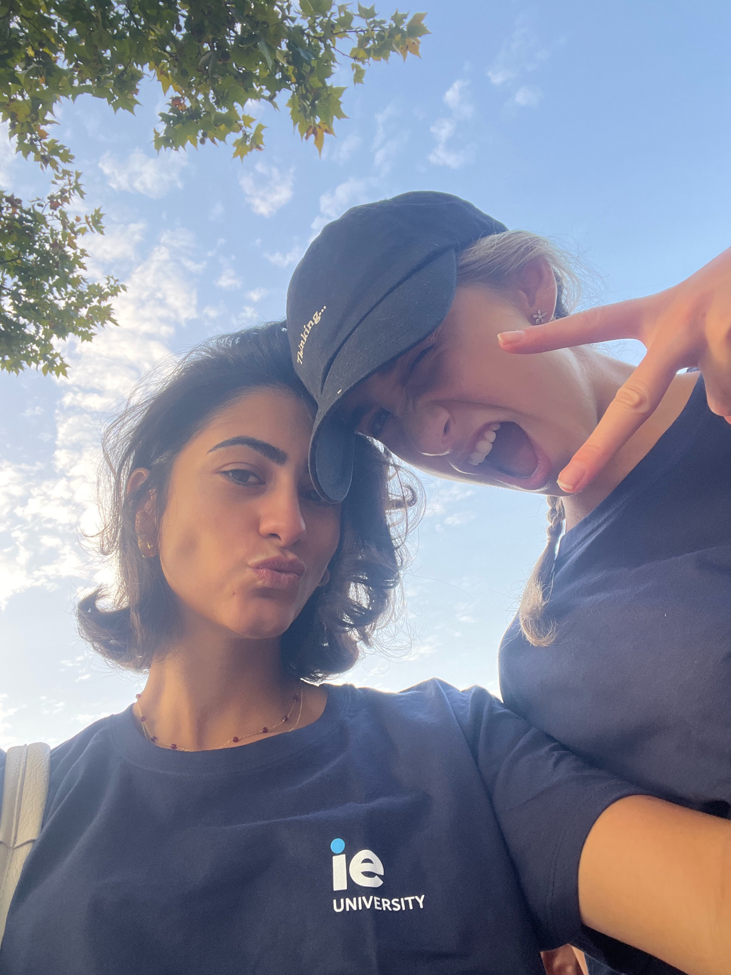 In Getafe, Spain, two young women are captured in a selfie outdoors, with a bright blue sky and scattered clouds forming the backdrop. A leafy tree branches into the upper left corner of the frame. Both individuals are wearing dark blue t-shirts with the "ie UNIVERSITY" logo in white on the front.
The woman on the left has dark, shoulder-length hair and is looking directly at the camera with her lips puckered. She is wearing a delicate gold necklace with small red beads. The woman on the right is wearing a black baseball cap and has blonde hair braided into a ponytail. She is leaning towards the left, with her mouth open in a laugh or shout and her right index finger extended, making a peace sign gesture towards the camera. A subtle earring is visible on her left ear. The perspective of the selfie is from a low angle, looking up at the subjects and the sky.