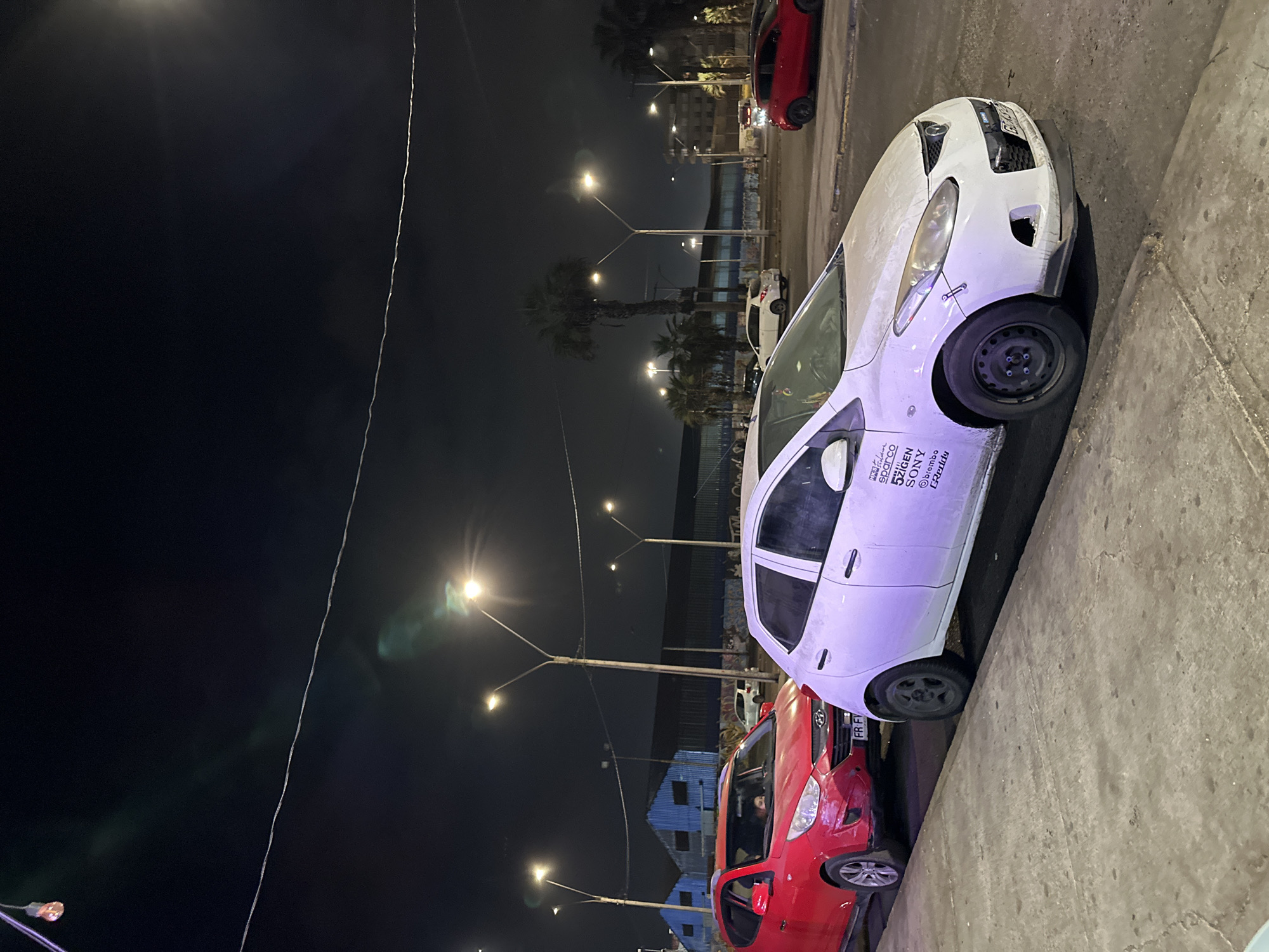 The image captures a street scene in Arica, Chile, at night. The primary focus is on parked cars along a curb. A white car, seemingly a Mazda 3, is parked close to the curb, appearing dusty and with visible decals on its side including "5zigen, Sparco, Sony, Brembo, and Eibach". Behind it is a red Hyundai i10 parked closer to the foreground. The surrounding area is illuminated by streetlights, and other vehicles are visible in the distance, including another red car at the far end of the street. Palm trees and buildings are present in the background. The overall environment suggests an urban setting with nighttime lighting.