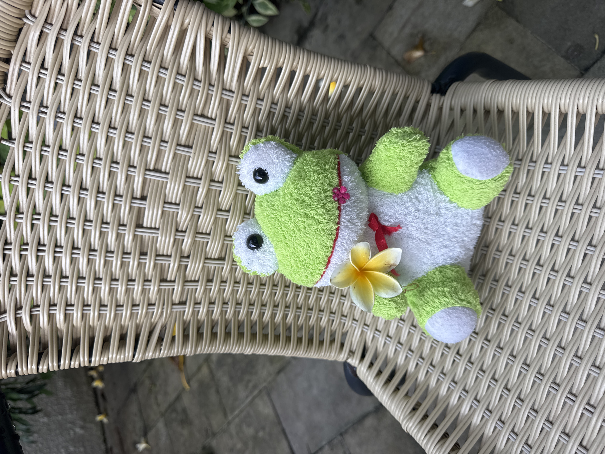 A green and white frog stuffed animal lies on its back on a woven chair in Kuta Selatan, Indonesia. The frog has a yellow flower placed near its head. The chair is positioned on a tiled surface, and green foliage is visible in the background.