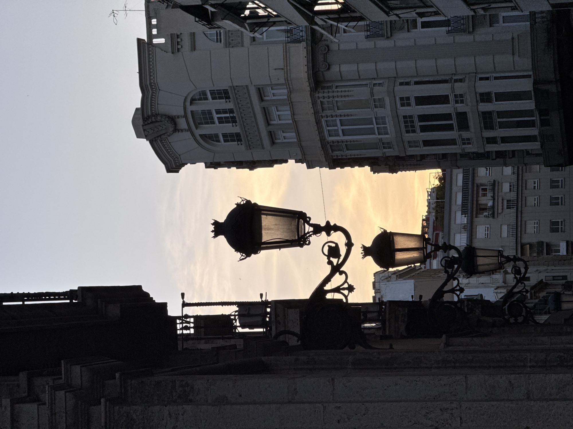 Captured in València, Spain, the image presents an architectural perspective looking upwards. Two vintage-style streetlights are silhouetted against a backdrop of soft, golden-hued sky, presumably during sunrise or sunset. The buildings flanking the sky's opening showcase classic European architecture, with detailed facades and numerous windows. The structures in the foreground are dark and undefined, creating a stark contrast with the brighter sky and buildings above.