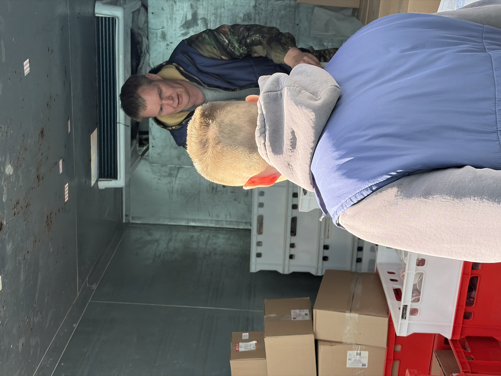 The image shows the interior of a delivery vehicle, potentially a refrigerated van, containing items such as cardboard boxes and red and white plastic crates. Two men are present. One man, with a darker complexion and wearing a jacket with a camo pattern, is closer to the front of the vehicle, partially obscured by the other man. The other man, with fair skin and short blond hair, is wearing a blue jacket and gray hoodie. The specific location of the scene within Chaikovsky, Russia cannot be determined from the image alone.