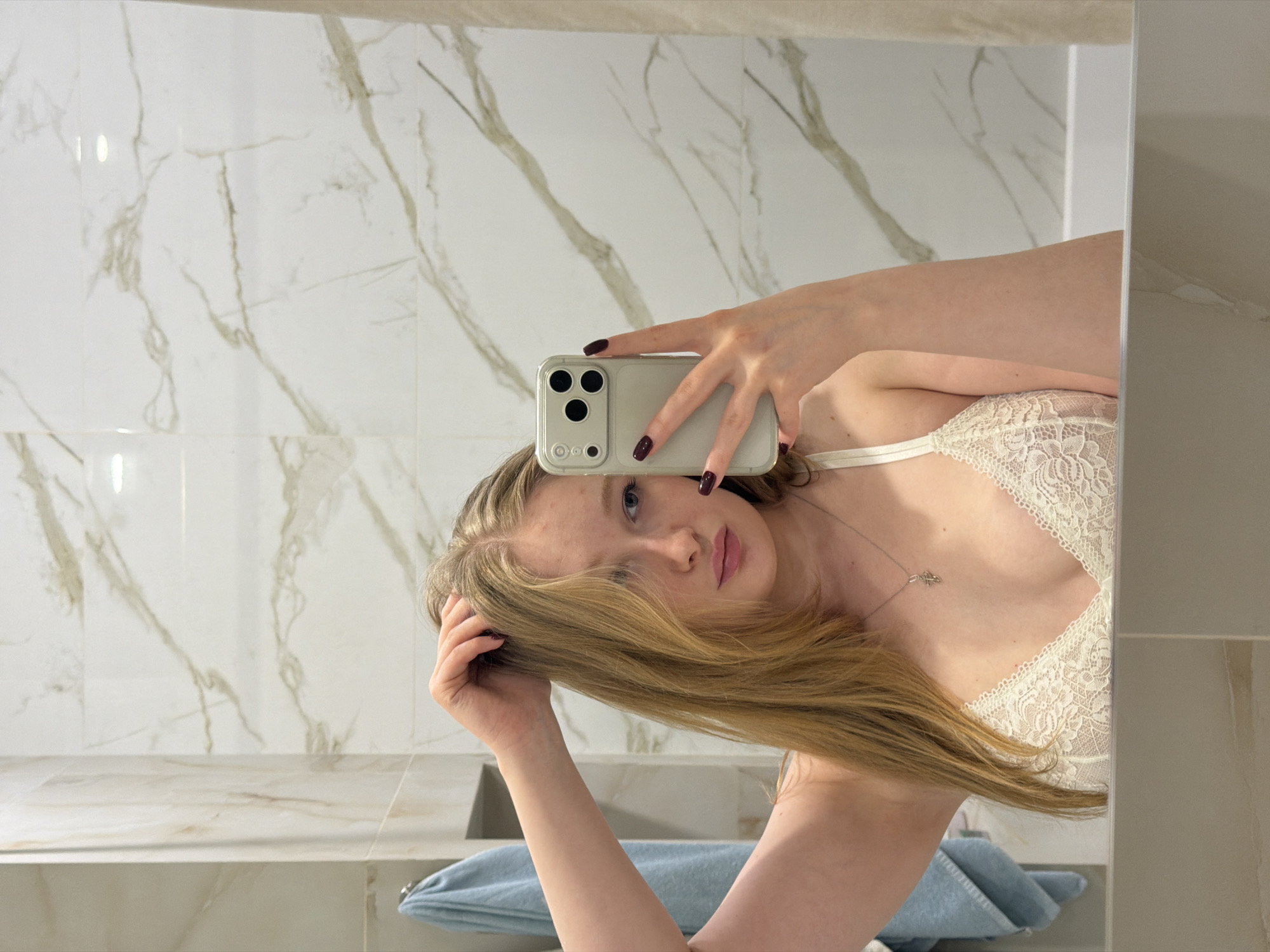 A young woman is taking a selfie in a bathroom. She is holding a phone with a triple-lens camera in her right hand. Her hair is blonde and she is wearing a white lace bra. A silver necklace is visible on her chest. A blue towel is present on the countertop. The bathroom has white marble walls. There is no indication that this image was taken in Rossiyskiy, Russia.