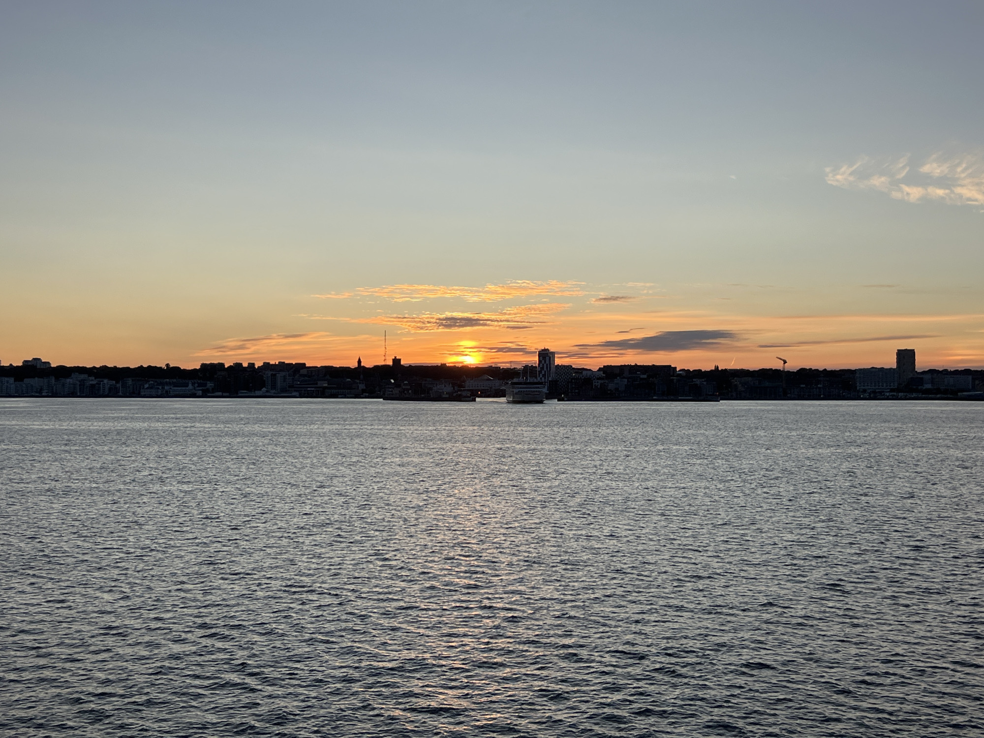 The image depicts a sunset over the Öresund strait, with the city of Helsingborg, Sweden, visible in the background. The sky is painted with hues of orange, yellow, and light blue, transitioning from the horizon upwards. Wispy clouds are scattered across the sky, catching the last rays of the sun. The sun itself is a bright, concentrated orb, partially obscured by clouds and the city skyline.
The water in the foreground is a dark, shimmering blue, reflecting the colors of the sky and the light from the setting sun. Gentle ripples disturb the surface, indicating a calm sea.
The Helsingborg skyline is silhouetted against the vibrant sky. Notable structures include a tall, modern building with a checkered facade on the right side of the frame, likely the Knutpunkten complex. A large ferry or cruise ship is visible in the middle distance, afloat on the water, its form softened by the twilight. To the left of the ferry, a crane stands as a stark silhouette against the sky, indicating ongoing construction or port activity. Further to the left, more buildings and structures form a continuous urban silhouette along the coastline. The overall impression is one of a serene evening in a coastal city.