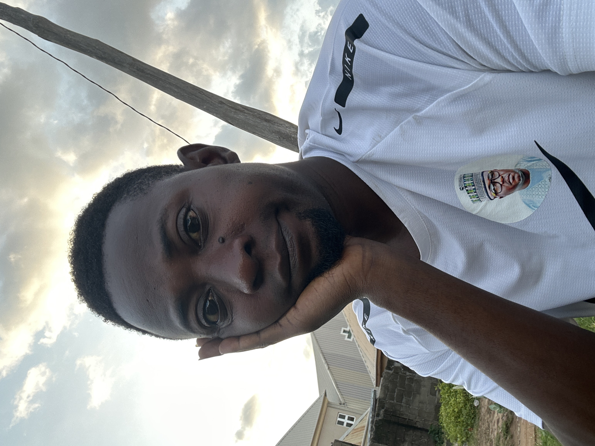 A medium shot selfie captures a Black man resting his head on his left hand, his eyes looking towards the camera. He has short, dark, curly hair and a goatee. He is wearing a white Nike t-shirt with black accents. A circular patch with a portrait of a man in traditional attire is visible on the left side of his chest. The background shows a cloudy sky, a wooden pole with a wire, and buildings with tiled roofs. This scene is located in the city of Narayi, Nigeria.