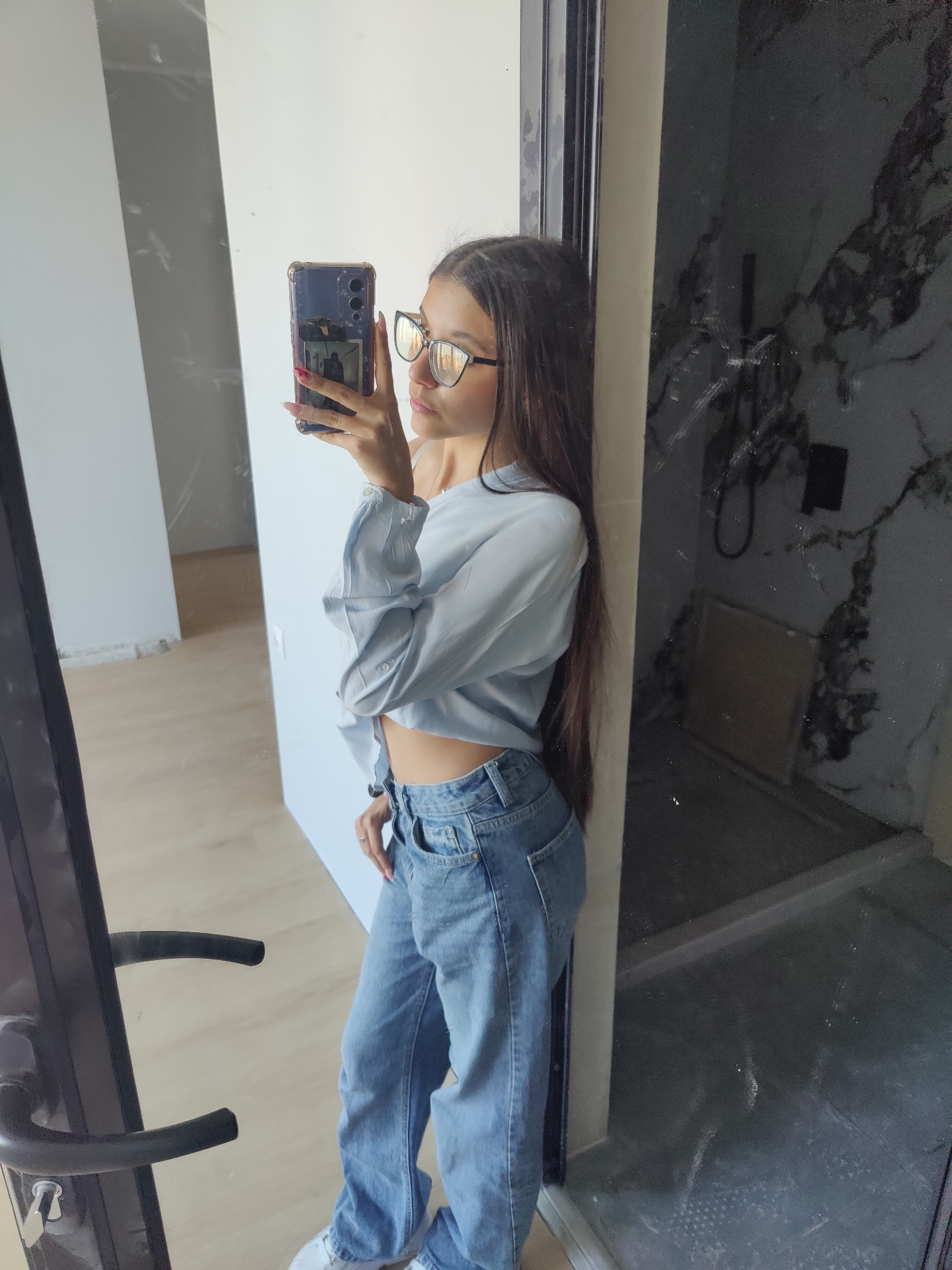 A young woman is taking a selfie in a room undergoing renovation. She is standing near a doorway. She is wearing glasses, a light blue blouse, and jeans. Her phone is in her right hand. The room behind her shows signs of construction, with unfinished walls and floors. The adjacent room has a grey marble design, likely a bathroom, with a shower visible. It is not possible to identify the exact location in Plovdiv, Bulgaria, based solely on this image.