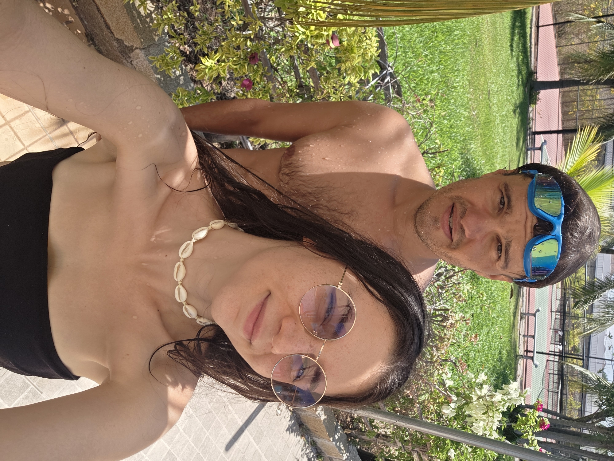 The image captures a selfie taken outdoors by a woman wearing a black top, shell necklace, and round sunglasses. Standing behind her is a man with wet hair and blue swimming goggles. The setting includes green grass, plants, and part of a building structure with a red roof. In the background, there is a net. The location is Phu Thuy Ward, Vietnam.