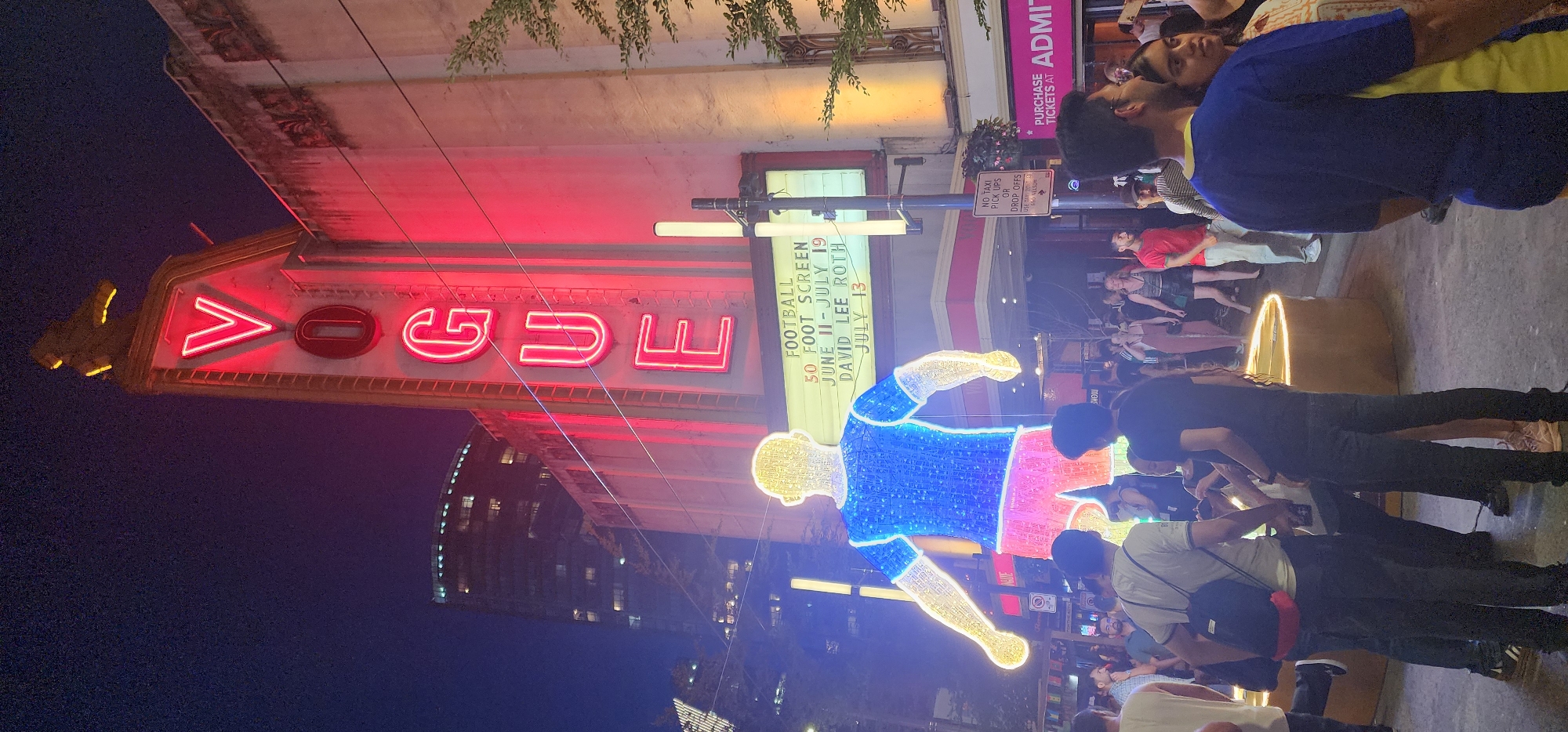 The image shows the exterior of the Vogue Theatre at night, with neon lights illuminating the marquee and building facade. A large, illuminated sculpture of a person in a soccer uniform stands in front of the theatre. A crowd of people are gathered on the sidewalk, some looking at the sculpture and others interacting with each other. The scene appears to be a lively event or gathering. The location is identified as Unknown, Unknown.