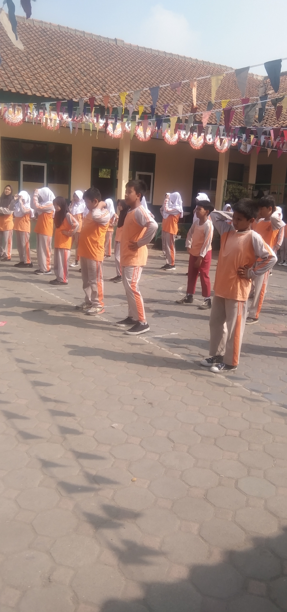 The image captures a group of children, predominantly young boys and girls, engaged in what appears to be a physical activity or exercise session outdoors. They are all wearing matching orange and grey uniforms, consisting of long-sleeved shirts and pants with orange stripes. Most of the children have their hands on their hips or are in a similar pose, suggesting they are following instructions. The background features a building with a tiled roof and decorative banners strung across. The ground is paved with interlocking hexagonal blocks, and shadows of the children and banners are visible on the pavement. The scene suggests a school or community event taking place in a sunny outdoor environment. While the specific location within the city of Rahayu, Indonesia is not explicitly stated, the architectural style and the presence of children in school uniforms are consistent with such a setting in Indonesia.