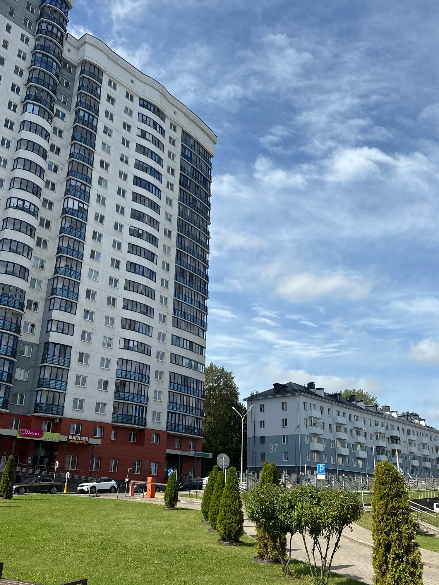 In Minsk, Belarus, an outdoor shot captures a modern residential complex under a partly cloudy sky. On the left, a tall, multi-story white building with blue-trimmed balconies and numerous windows dominates the frame. To the right, a series of lower, light blue apartment buildings with darker blue trim stretch into the distance. In front of the buildings, a well-maintained green lawn leads to a paved walkway. Several vehicles, including a white SUV, a taxi, and a dark car, are parked near a barrier and a building entrance, where signs display Russian text. Trees and manicured bushes are strategically placed throughout the landscape. The scene appears to be a calm, urban residential area.