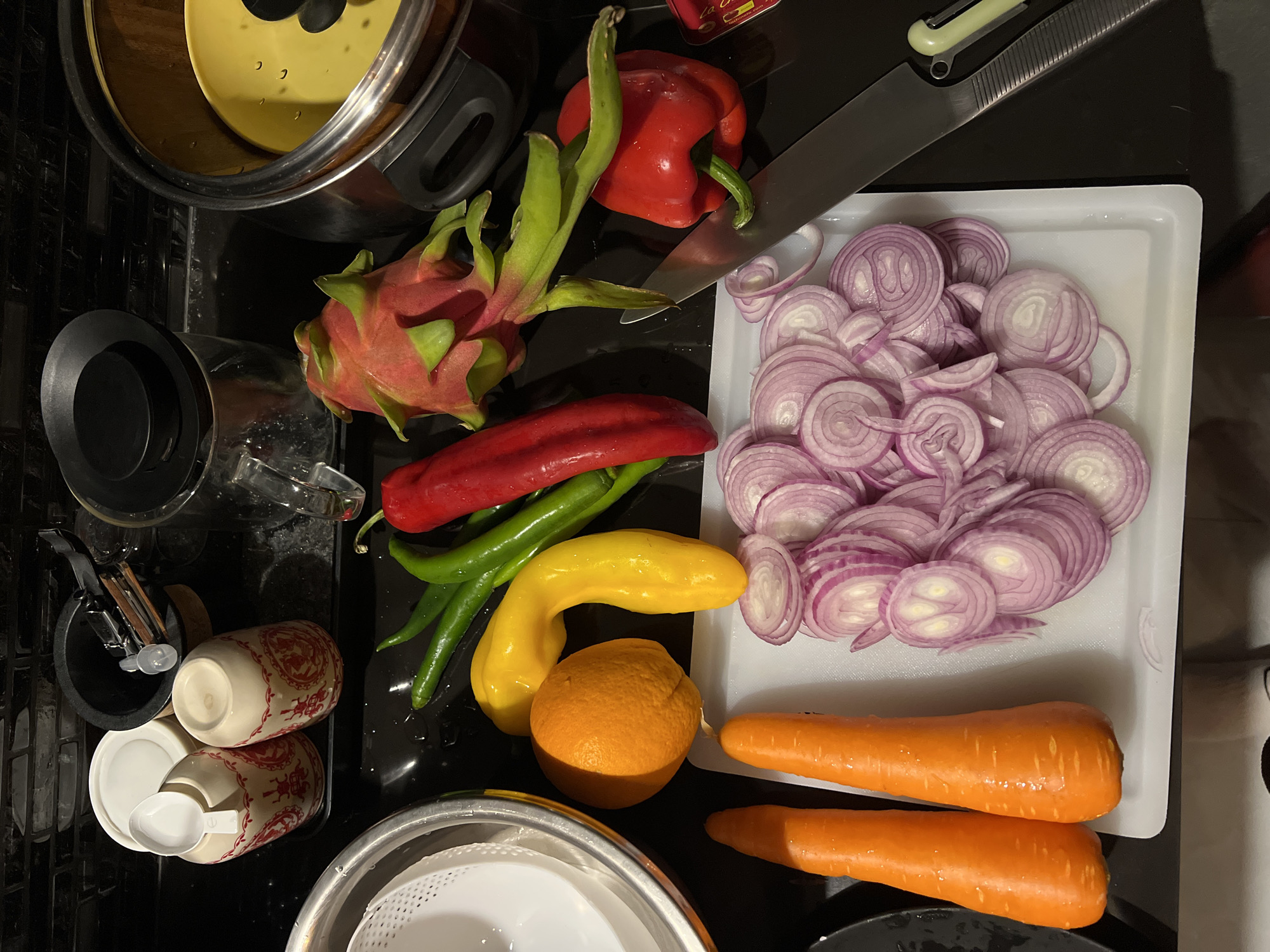 The image depicts a kitchen scene with various ingredients and utensils on a black countertop. A stainless steel pot and glass pitcher are on the left. A red bell pepper, dragon fruit, and long red chili pepper lie near a white cutting board. The board holds a pile of sliced red onions. A yellow bell pepper and orange are near two carrots. A knife is on the right side of the bell pepper, above the cutting board. Below the pitcher, there is a spice container with a spoon, a coffee grinder, and a strainer. The location is a kitchen, but the city and specific location are unknown.