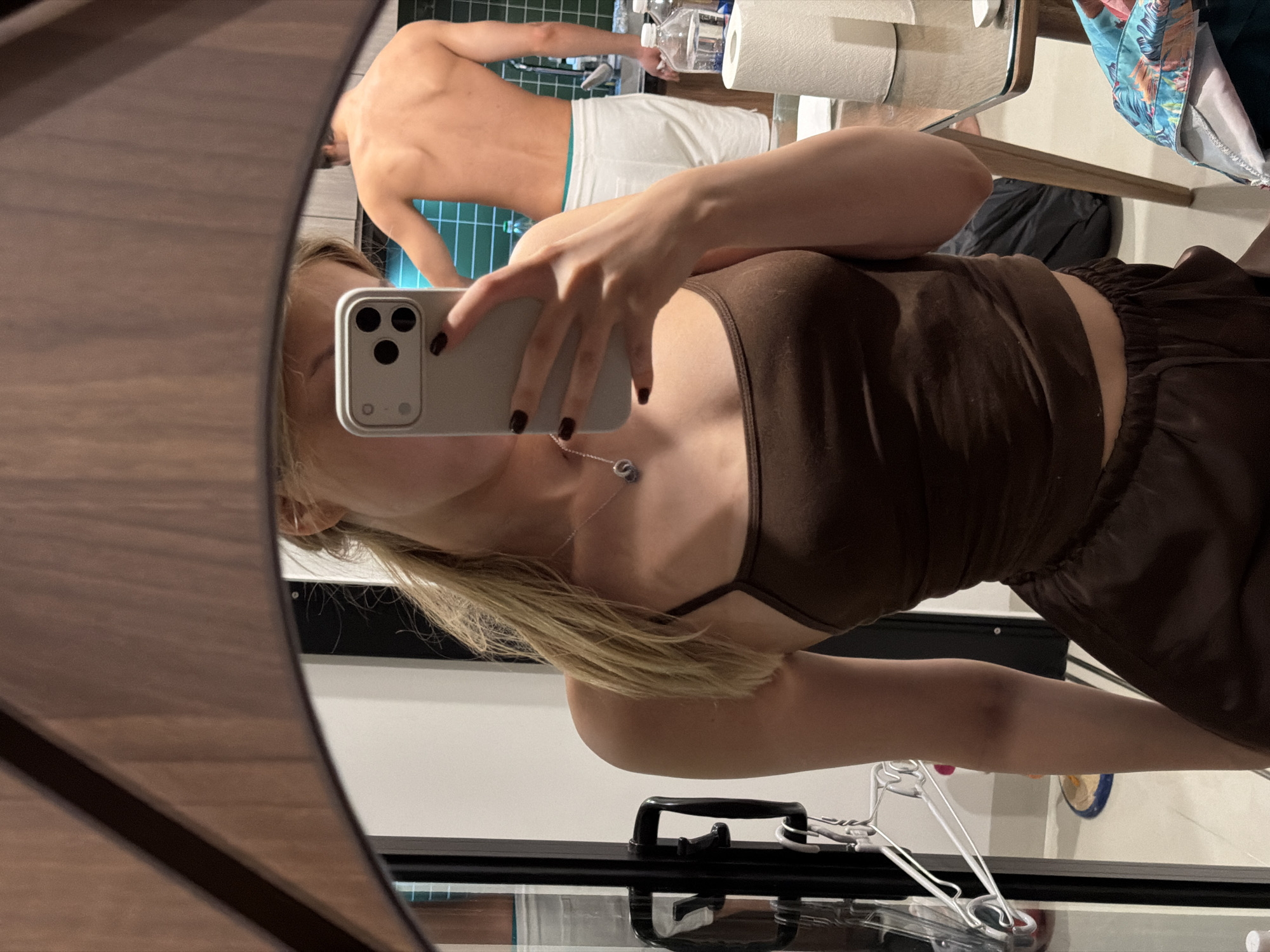 The image depicts a woman taking a mirror selfie in what appears to be a home bathroom or dressing area. She is wearing a brown camisole and brown shorts. A silver necklace with a circular pendant is visible. Her hair is blonde. In the mirror's reflection, a person with their back to the camera is visible, wearing white shorts. Behind them, there are toiletries including what looks like a roll of toilet paper and a plastic bottle. The lighting suggests it could be daytime or artificial indoor lighting. The overall scene is casual and personal.