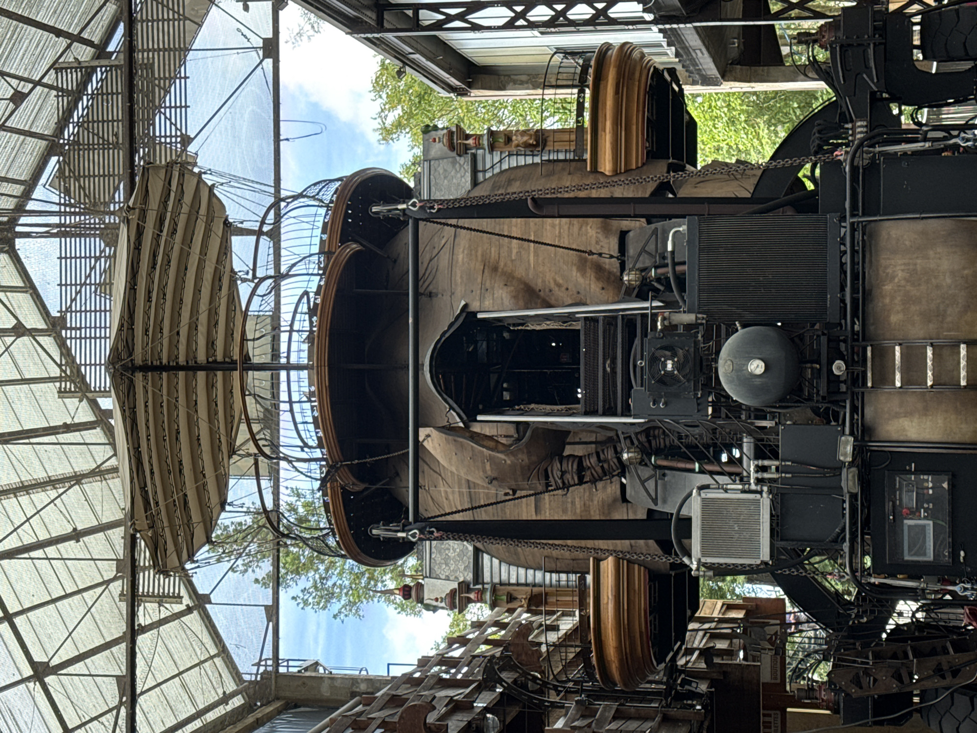 This is a full-frame, eye-level, medium shot of a large, steampunk-inspired mechanical structure resembling a gigantic elephant head, housed within a greenhouse-like structure. The structure is made of dark wood and metal, with intricate details like chains, pipes, and mechanical components visible. The elephant's head has large, wooden tusks and a small, dark entranceway serving as its mouth. Above the head, a tiered platform with a circular railing is suspended by chains, topped with a striped fabric canopy. The entire structure is set against a backdrop of lush green trees and a blue sky, visible through the transparent roof and walls of the greenhouse. The scene is located at Les Machines de l'Île in Nantes, France, a mechanical art park. There are no people visible in the image.