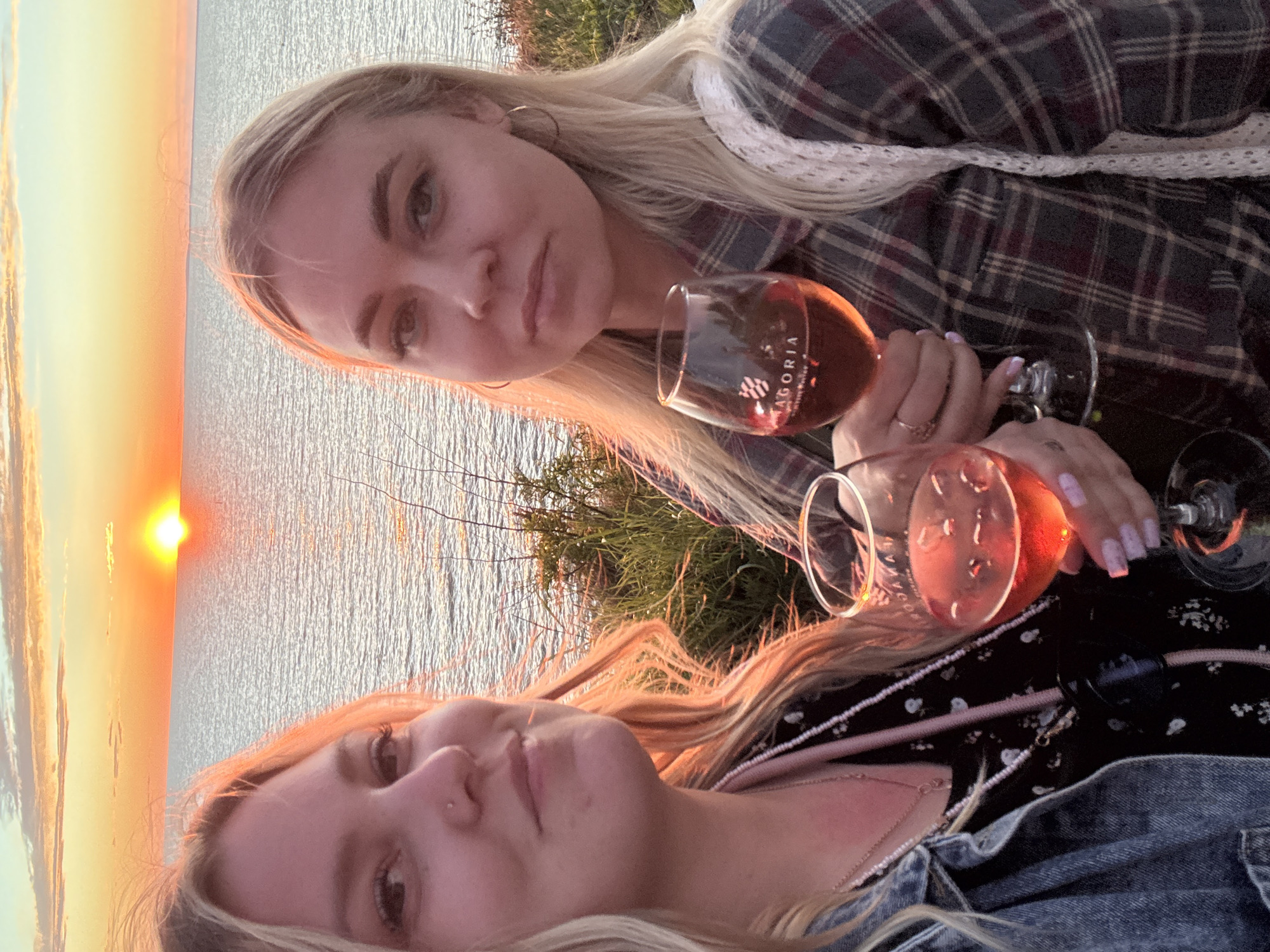 The image, originally rotated 90 degrees clockwise, captures two young blonde women enjoying an outdoor sunset by a large body of water, likely near Starotitarovskaya, Russia. The sun is visible as a bright orange orb positioned on the horizon, casting warm, golden light across the scene and reflecting vibrantly on the rippled water surface. Green foliage and small bushes line the elevated land separating the women from the water.
The woman in the upper part of the frame wears a dark plaid shirt and hoop earrings. She holds a stemware glass containing a rosé-colored beverage, with the brand name "AGORIA" and additional text visible on the glass. The woman in the lower part of the frame is seen wearing a dark top with a light floral pattern and a denim jacket. Her hand, adorned with light pink manicured nails, is near another identical glass of rosé-colored drink. Both individuals appear relaxed, savoring the scenic view and the tranquil atmosphere of the sunset.