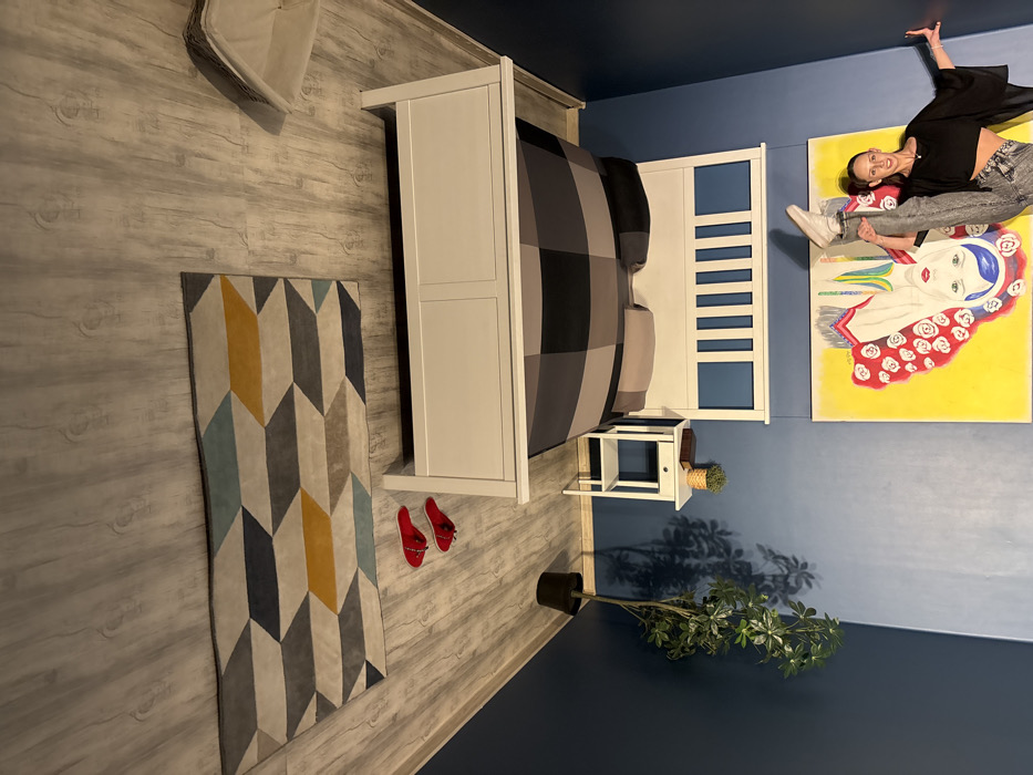 This image depicts an intentionally inverted bedroom scene within the Upside Down House attraction in Sofia, Bulgaria. A single adult female subject is shown mid-action, performing a high vertical leg kick with her right leg, appearing to stand on a dark blue wall. She wears a black cropped top, high-waisted grey patterned jeans, and white sneakers, smiling at the camera.
The room's design creates an optical illusion of being inverted. The actual ceiling, adorned with light wood-grain patterned wallpaper, is arranged to appear as the floor. Mounted inverted to this "floor" are several items: a white wooden bed frame with a grey and black checkered comforter and two grey pillows; a white wooden nightstand featuring a small potted plant in a woven basket; a larger potted plant with green foliage in a black pot; a rectangular rug with a geometric pattern in grey, blue, and yellow; and a pair of red flat shoes. A large rectangular artwork with a bright yellow background and a stylized, possibly inverted, face with red roses is affixed to the dark blue wall behind the subject.