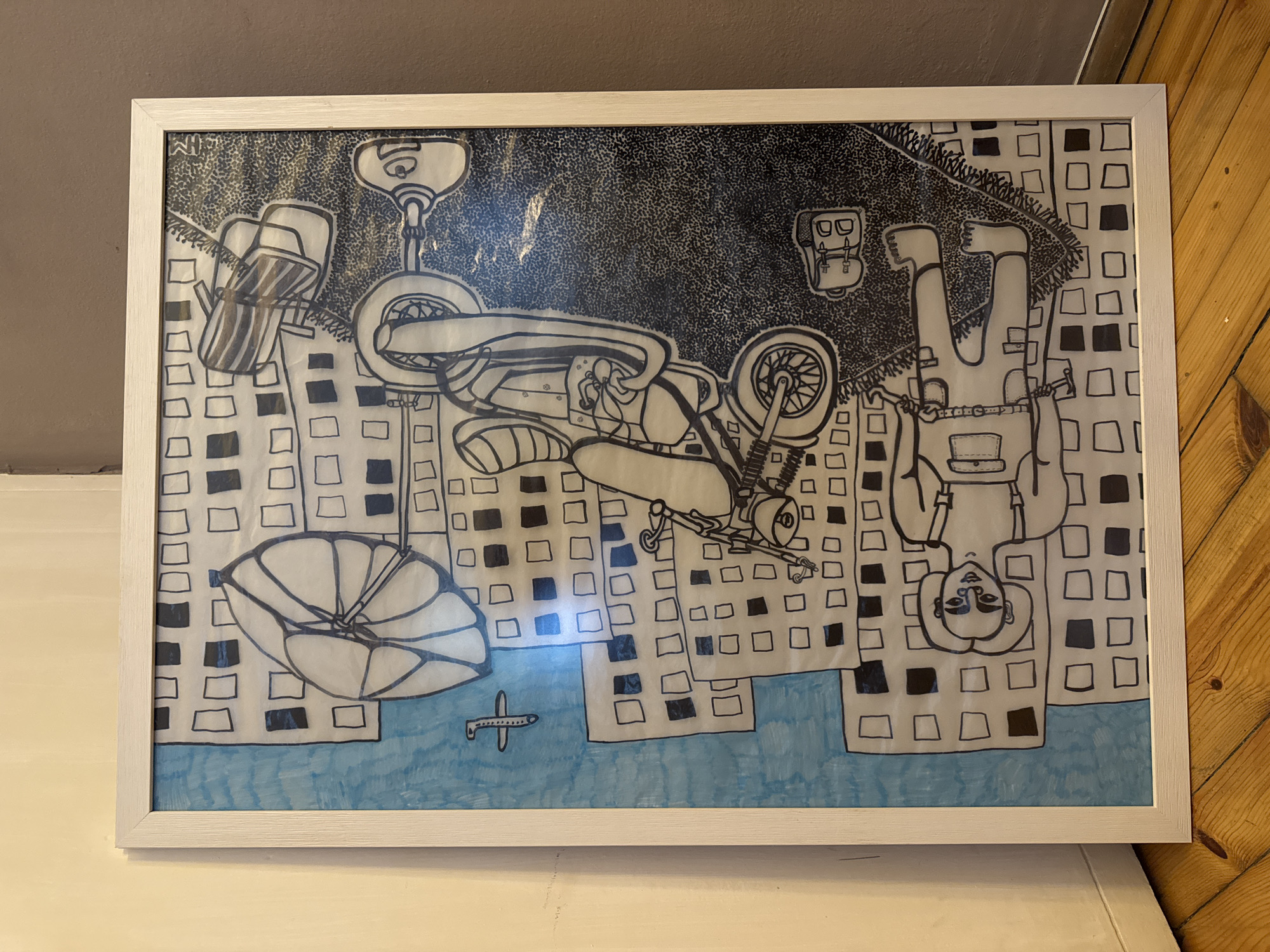 A framed, stylized drawing depicts an inverted urban scene. The central subjects are a person and a motorcycle, both appearing upside down and seemingly flying or falling over a cityscape. The person is situated on the right side of the frame, head downwards, wearing a backpack and what looks like a belt with pouches. The motorcycle is positioned above the person, also inverted, showing its wheels, handlebars, and engine details.
Below the main subjects, an abstract city with numerous buildings is illustrated, primarily using outlines of squares and rectangles, some filled in black to create a patterned effect. Towards the bottom left, an upside-down parachute or paraglider is visible. A body of blue water extends across the lower portion of the drawing, upon which a small, simple outline of an airplane is depicted. The sky or background above the city is filled with a dense, stippled pattern, creating a dark, textured effect. The artwork is contained within a light-colored frame. No discernible text is visible within the drawing.