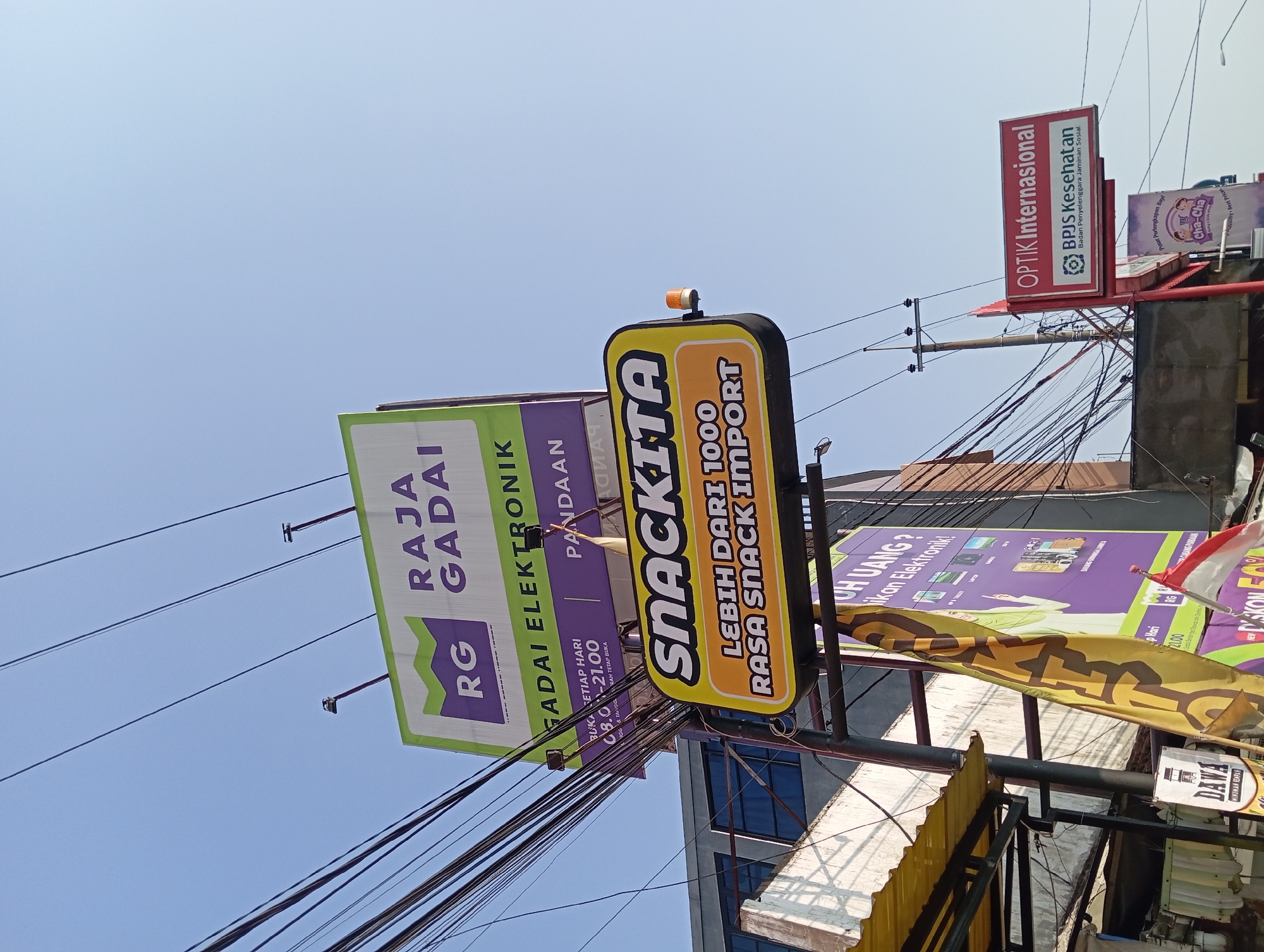 The image depicts an outdoor scene with several signs and a clear blue sky. The dominant signs are for "RAJAGADAI ELEKTRONIK" and "SNACKKITA." The "RAJAGADAI ELEKTRONIK" sign is a large billboard with green and white branding, featuring the initials "RG" in a purple graphic. Below this, a smaller sign indicates business hours from 08:00 to 21:00, open daily. The "SNACKKITA" sign is a bright yellow, oval-shaped sign with bold white lettering, advertising "LEBIH DARI 1000 RASA SNACK IMPORT." There are numerous electrical wires crisscrossing the frame, suggesting an urban or semi-urban environment. To the right, a smaller red sign reads "OPTIK Internasional" and below that, "BPJS Kesehatan." Further down, another sign with a purple background has graphics of electronic items and text in Indonesian. A yellow flag with black lettering is partially visible on the left. The image appears to be taken from a low angle, looking upwards towards the sky and the signs. The overall impression is of a commercial street in Petungasri, Indonesia, with various businesses advertising their products and services.