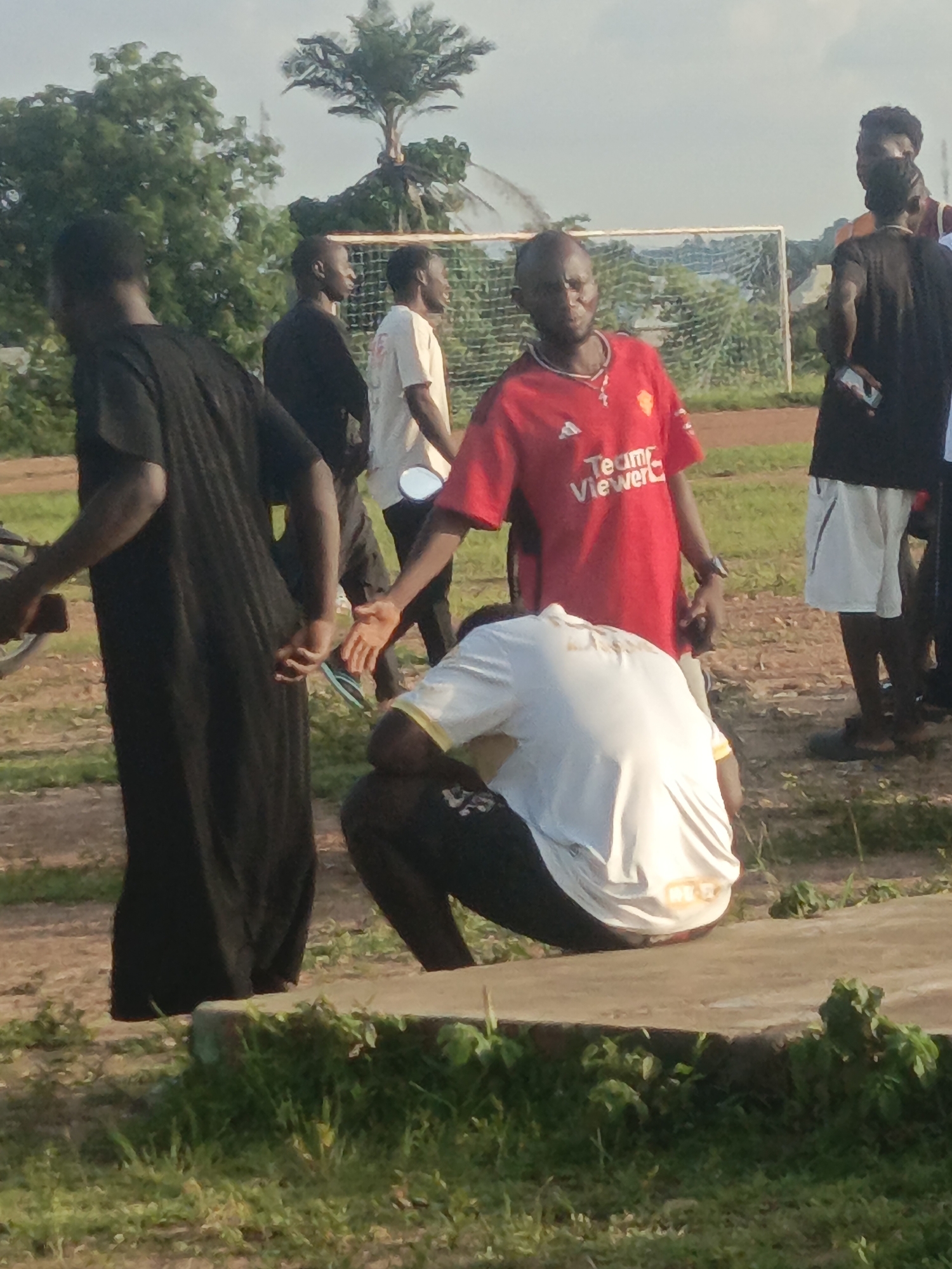 In Akwanga, Nigeria, at a soccer field, several individuals are present. The foreground is dominated by a man in a red Manchester United jersey, holding hands with another person. To the left, a man in a long black robe is seen. In the background, more individuals are visible, along with a soccer goal. The environment suggests an outdoor setting, possibly during daylight hours, with greenery and a light sky.