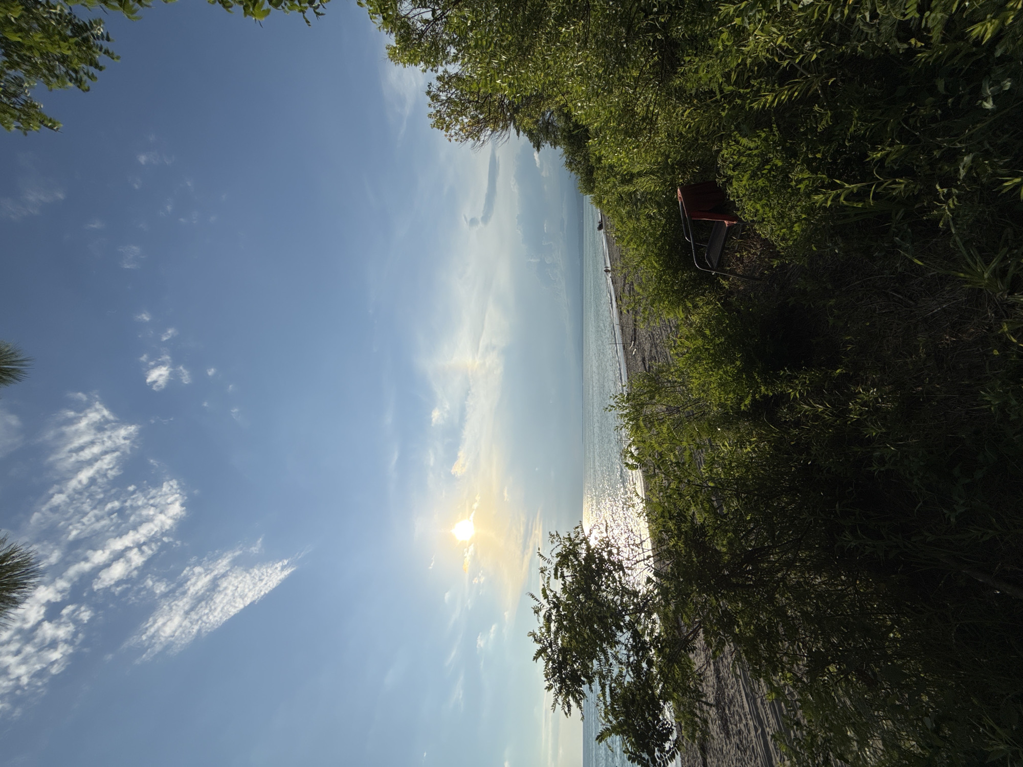 The image depicts a serene beach scene at Yuada-Gulrypsh, Abkhazia. The sun, low in the sky, casts a warm glow on the water, creating a shimmering effect on the surface. Wispy clouds streak across the vast blue sky, with a hint of a rainbow visible in the distance. Lush green foliage frames the foreground, with a beach and calm ocean stretching out to the horizon. A solitary red chair rests amidst the greenery, adding a touch of color to the natural landscape. A few distant figures are visible on the beach, suggesting a tranquil day by the sea.