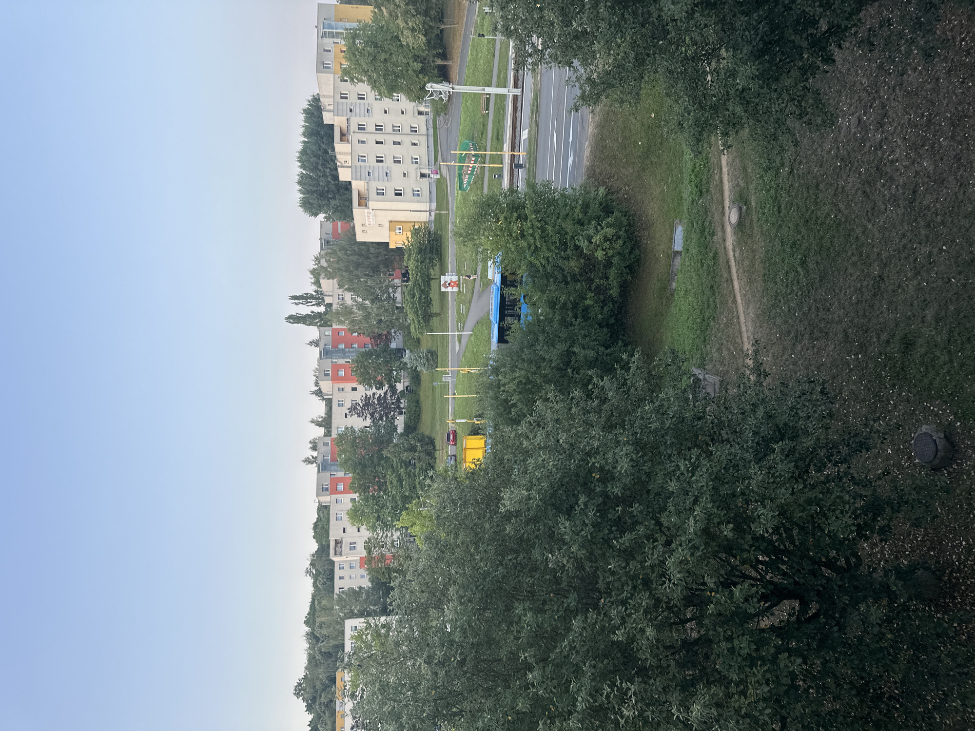 This is an eye-level, medium shot of a residential area in Unknown, Unknown, under a clear, pale blue sky. In the foreground, dense green trees with a slightly dark hue dominate the left and right sides of the frame, partially obscuring the view of the middle ground. Beyond the trees, a row of apartment buildings stretches across the horizon. These buildings are varied in color, with sections of gray, white, red, and yellow. Some buildings have visible windows and balconies.
In the middle ground, a road runs horizontally. A bright blue bus is in motion on this road, positioned in front of some trees and buildings. To the left of the road, there is a pedestrian path with a few people visible, one walking and another standing near a colorful, possibly artistic, display. There are also yellow poles and some green fencing elements scattered throughout this area. Further back, more apartment buildings and trees are visible, with a subtle incline to the terrain. The overall impression is a suburban or residential district with a mix of green spaces and multi-story housing.