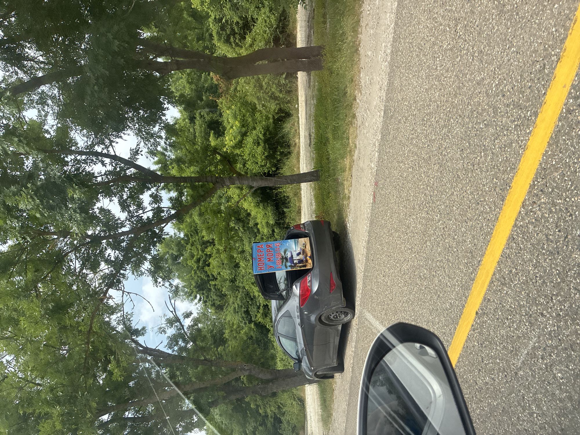 An eye-level, medium shot captures a gray sedan parked on the side of a road with its trunk open. A blue advertisement sign is propped in the trunk, displaying text in Russian and an image of a beach scene. The sign reads "НОМЕРА У МОРЯ" which translates to "ROOMS BY THE SEA" and a phone number "8-988-588-74-45". The car is positioned on a gravel and dirt shoulder next to a paved road with a yellow line. Lush green trees and foliage line the background. The sky is visible through the trees, with some clouds present. The scene appears to be in Arkhipo-Osipovka, Russia, a resort town known for its coastal location. The perspective suggests the photo was taken from another vehicle traveling on the road, as a car's side mirror is visible in the lower left corner of the frame.