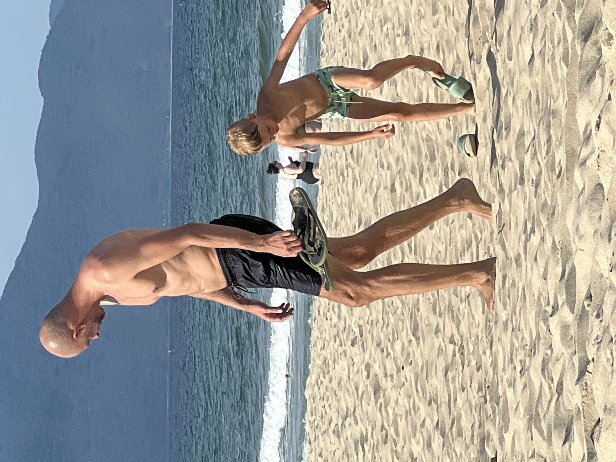 A man and a boy walk on a sandy beach with the ocean and mountains in the background. The man, who is bald and shirtless, wears black swim trunks and carries a pair of flip-flops in his right hand. He is looking down. The boy, who is shirtless and wears green patterned swim trunks, is bending down and picking up a pair of green flip-flops with his right hand. In the background, other beachgoers can be seen near the water. The sky is clear and blue, and the water is a deep blue with waves breaking on the shore. The mountains in the distance are a muted blue and appear to be covered in trees. This scene is consistent with a location such as My Khe Beach in Da Nang, Vietnam, known for its wide sandy shores and backdrop of the Son Tra Peninsula mountains.