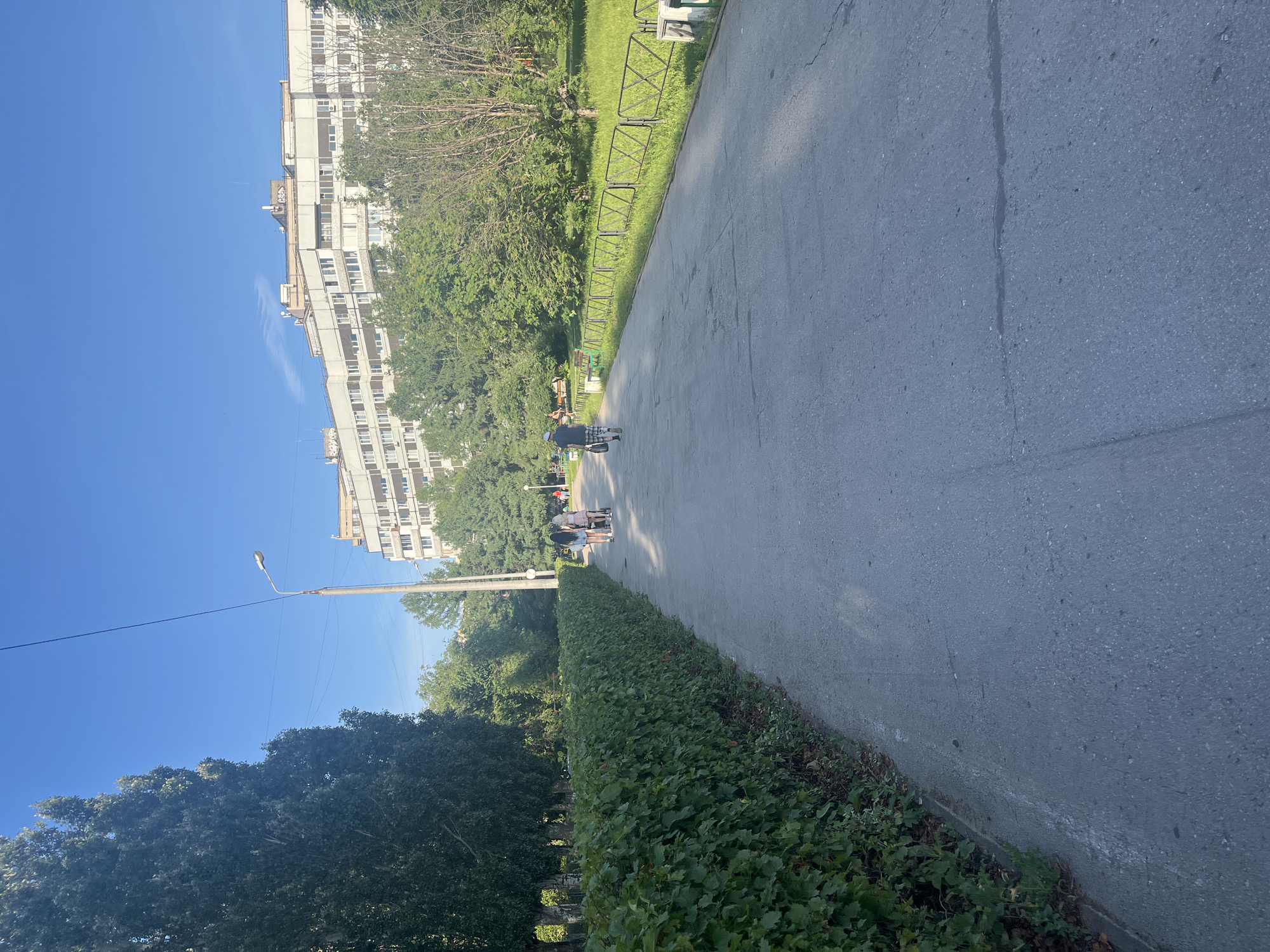 The image depicts a sunlit outdoor scene, possibly a park or a tree-lined street, with a clear blue sky. On the left side of the frame, a tall, multi-story building with many windows is visible, flanked by lush green trees and foliage. A sidewalk or path runs diagonally through the center of the image, with a neatly trimmed hedge bordering it on the right.
Several people are present, walking along the path. One individual, wearing a dark shirt and light-colored shorts, is seen from behind, walking away from the camera. Further down the path, a group of people, possibly a family or friends, are also walking together. One person appears to be pushing a stroller or a cart.
A lamppost stands to the left of the path, its light fixture silhouetted against the bright sky. A thin wire stretches from the lamppost across the frame. The ground is paved with asphalt, showing some cracks and wear. The overall atmosphere appears peaceful and leisurely, suggesting it's a pleasant day for an outdoor stroll. The sunlight casts shadows, indicating it is likely midday. There is no visible text in the image.