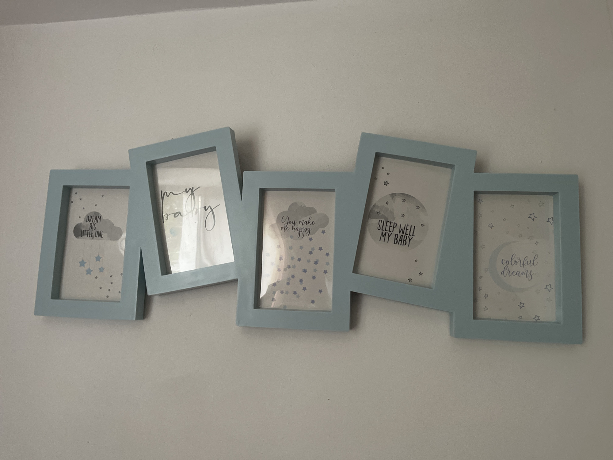 A pastel blue, multi-photo frame is mounted on a white wall. The frame is arranged in an angled, staggered fashion, with five individual rectangular openings. Each opening contains a printed image with text. From left to right, the images depict: a cloud with stars and the text "DREAM BIG LITTLE ONE"; a cursive phrase "my baby"; a cloud with stars and the text "You make me happy"; a moon with stars and the text "SLEEP WELL MY BABY"; and a crescent moon with stars and the text "colorful dreams". The wall appears to be a plain, untextured surface. There are no people or animals visible in the image. The overall scene suggests a nursery or child's room decoration. The location is identified as Kohtla-Järve linn, Estonia.