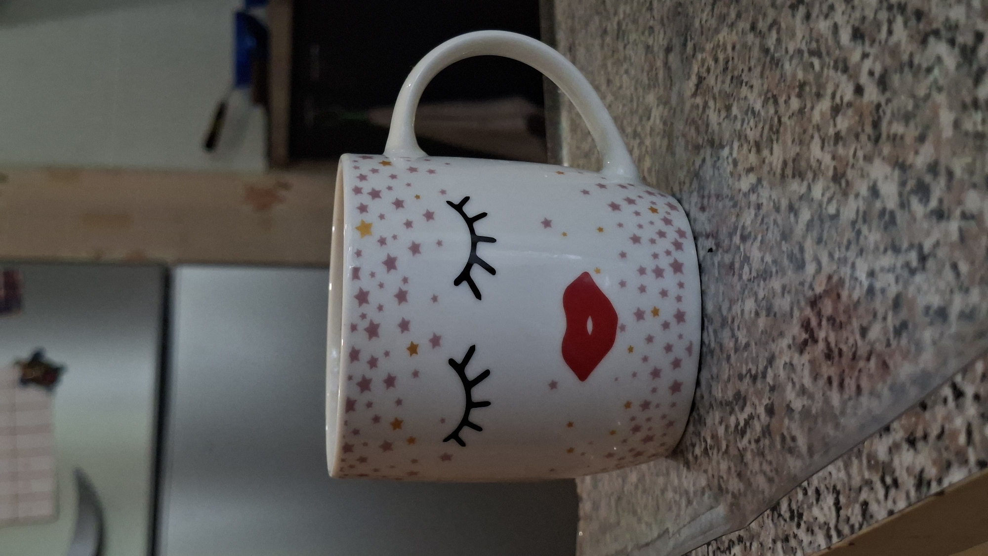 A white mug with closed eyes and red lips design, decorated with pink and yellow stars, sits on a speckled granite countertop. In the background, a stainless steel refrigerator is visible. The scene is an indoor environment, likely a kitchen. The image does not provide any information about the city of Myadzyel, Belarus.