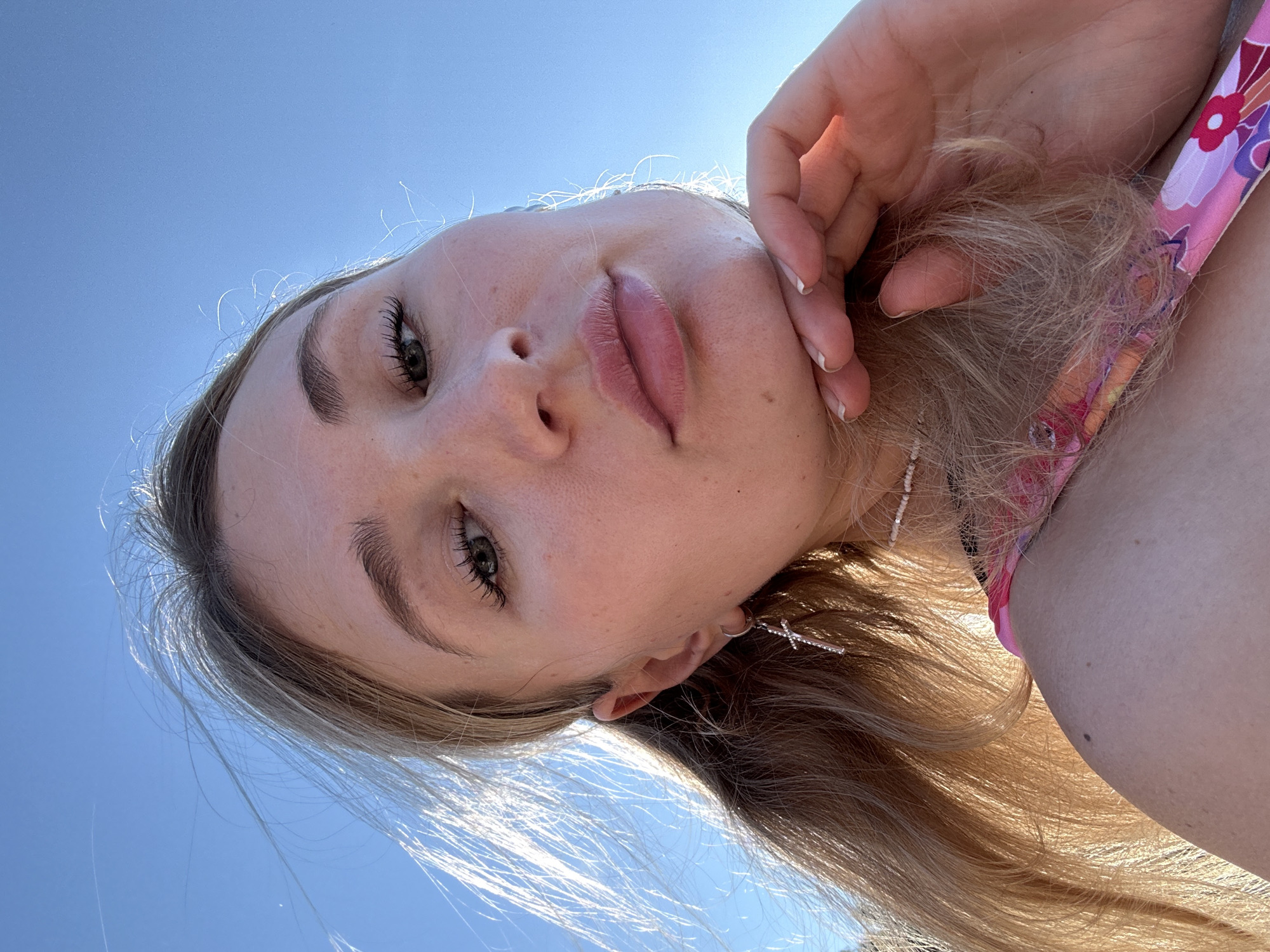 An outdoor, close-up, waist-up selfie shows a young Caucasian woman with blonde hair pulled into a ponytail against a bright blue sky. She is wearing a pink and floral patterned top and a silver cross earring with rhinestones. Her left hand is resting on her chin, and her hair is blowing in the wind. The image appears to have been taken in the city of Chaikovsky, Russia, given the angle and clarity of the sky.