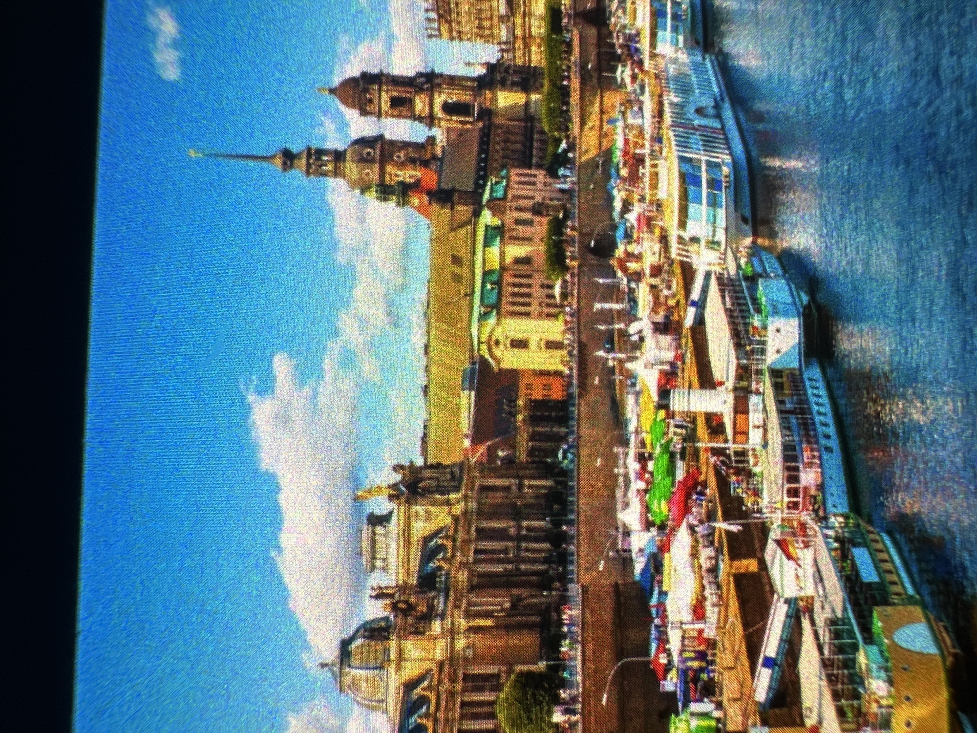 The image depicts a waterfront scene with architectural landmarks and several boats docked in a body of water. In the background, a large, ornate building with domes and spires dominates the skyline, suggesting a historical or cultural significance. In the foreground, a lively market or festival is set up along the waterfront, with numerous tents and stalls displaying various goods. Several boats, including passenger ferries and smaller vessels, are moored along the quay, some with people visible on deck. The water appears calm, reflecting the blue sky and the surrounding structures. The overall atmosphere is one of a bustling urban environment with a blend of historical architecture and contemporary activity.