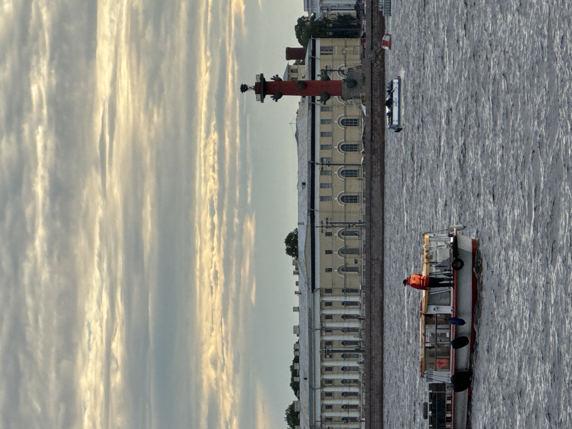 This is a photograph of the Neva River in Saint Petersburg, Russia, taken during a cloudy sunset. In the foreground, a white and red boat with a glass-enclosed cabin is moving on the water. A person in an orange life jacket stands on the deck of this boat. In the middle ground, a red rostral column, a historical landmark, stands on the Spit of Vasilyevsky Island. Behind the column is a long, light-colored neoclassical building with a colonnade. Further in the background, on the right, a white motorboat with several passengers is visible. The water is choppy, and the sky is filled with layered clouds illuminated by the setting sun.