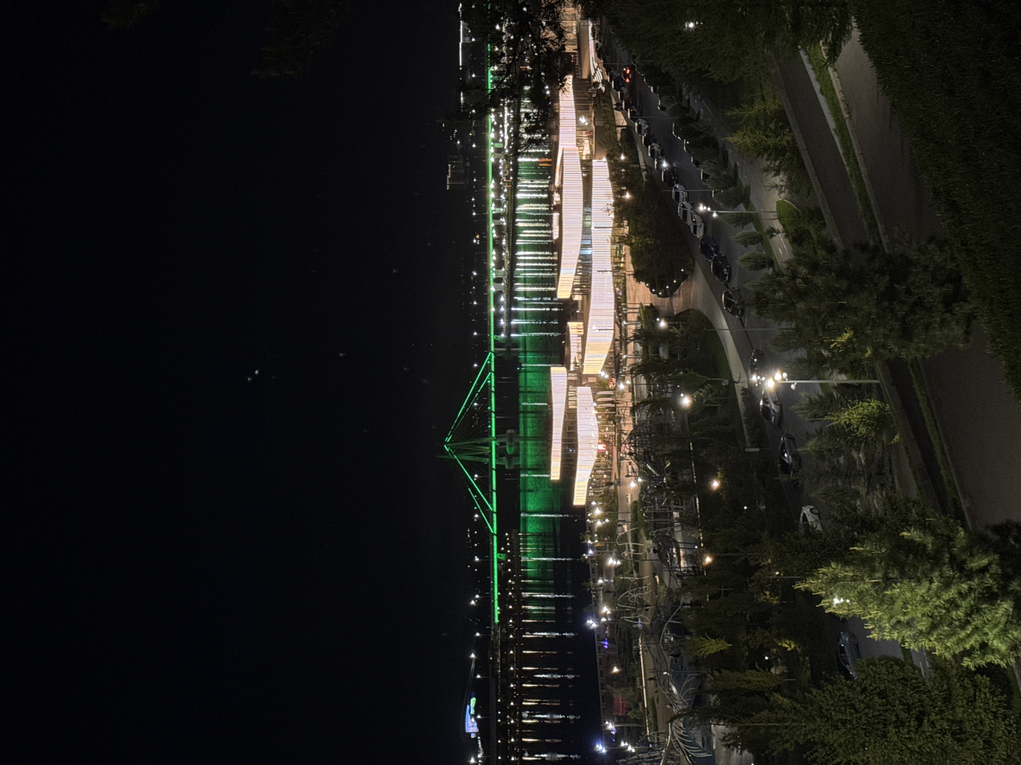 The image depicts a nighttime view in Kazan, Russia, featuring the Millennium Bridge illuminated in green. The bridge spans a body of water reflecting the bridge's lights and other illuminated structures. Adjacent to the water, a boardwalk is lit with white lights. A street with parked cars is visible along the boardwalk. The area includes trees and vegetation. The sky is dark.