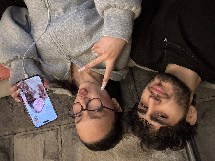 An indoor scene depicts two individuals, a male and a female, positioned closely together and looking towards the camera. The male, on the left, has dark curly hair, a beard, and wears a black long-sleeved shirt with a silver chain and small pendant; he has a slight smile. The female, on the right, has dark hair pulled back, wears eyeglasses with dark frames, and a grey zippered hoodie. Her lips are pursed in a pout, and her left hand is raised, forming a 'peace' sign. Her right hand holds a dark-colored smartphone, possibly an iPhone, displaying a video call or photograph of a third female with long brown hair, smiling and wearing headphones. A white charging cable is connected to the bottom of the smartphone. The background features a wall constructed from stacked, irregularly shaped grey and brown stone blocks. A hint of an orange object is visible in the lower right background. The specific location within Yerevan, Armenia, cannot be determined from the visual evidence in the image.