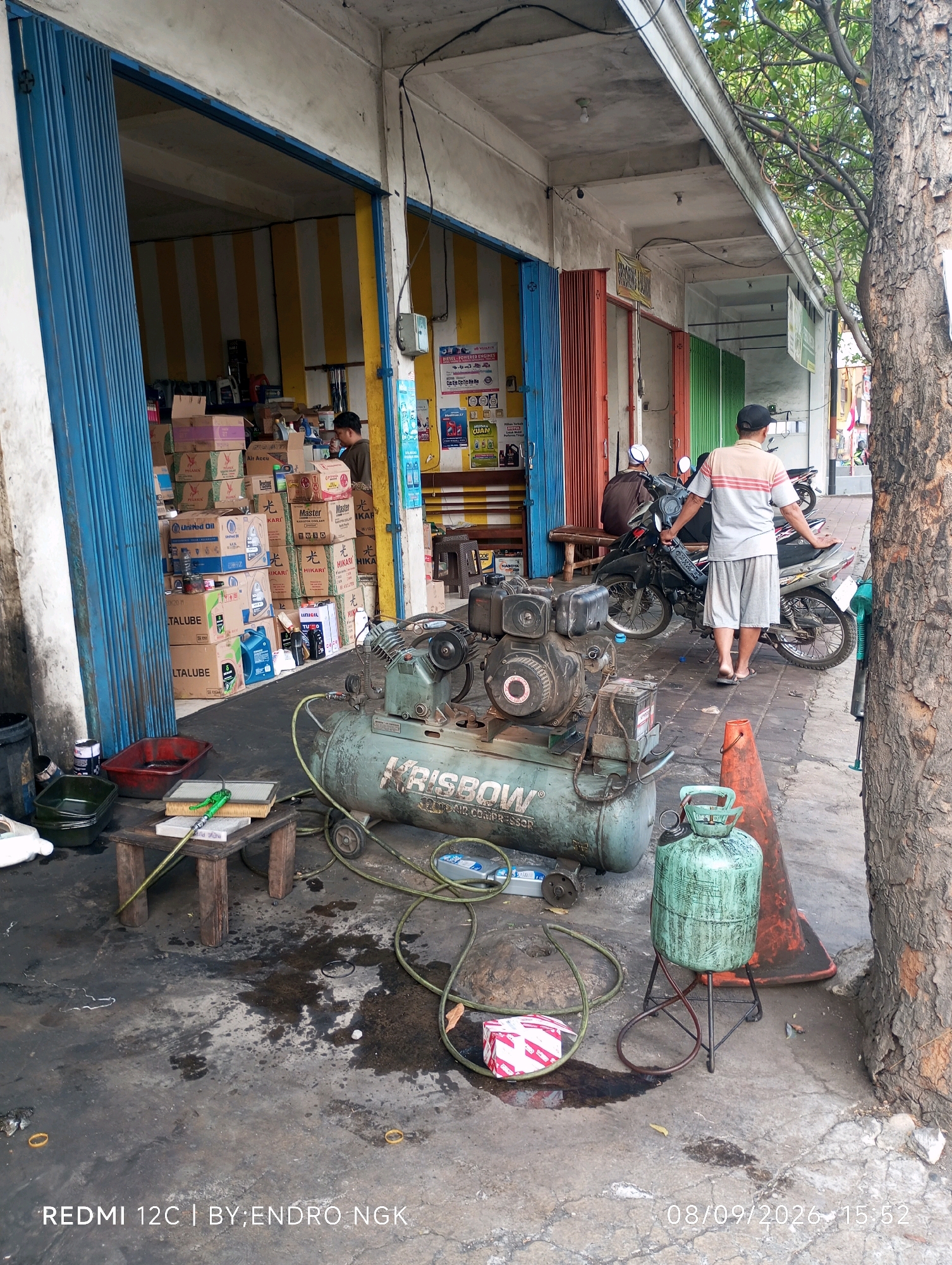 The image depicts a street-level view of what appears to be an automotive repair shop or a related business in Ringinanom, Indonesia. In the foreground, a large, green air compressor with the brand name "RISBOW" is prominently positioned on a paved surface that shows signs of oil stains and dirt. Adjacent to the compressor, a green gas cylinder with a metallic stand is visible. A small wooden stool with an air filter and a green hose is situated to the left of the compressor. Further left, there are several plastic containers, one red and one dark green, and a white object that might be a part of a cleaning supply or tool.
In the background, a man wearing a striped t-shirt and grey shorts stands near several motorcycles. He is facing away from the camera. Behind him, the open entrance of a shop is visible, revealing stacked boxes of various sizes, many with brand names like "PEGANUS" and "Master." The shop interior is lined with vertical yellow and white striped curtains. To the right of the man and motorcycles, a large tree trunk is present, and part of a building with orange and green roller doors is visible. The overall environment suggests an outdoor, commercial setting in a potentially busy area. The timestamp "08/09/2026 15:52" is overlaid on the image, along with "REDMI 12C | BY:ENDRO NGK".