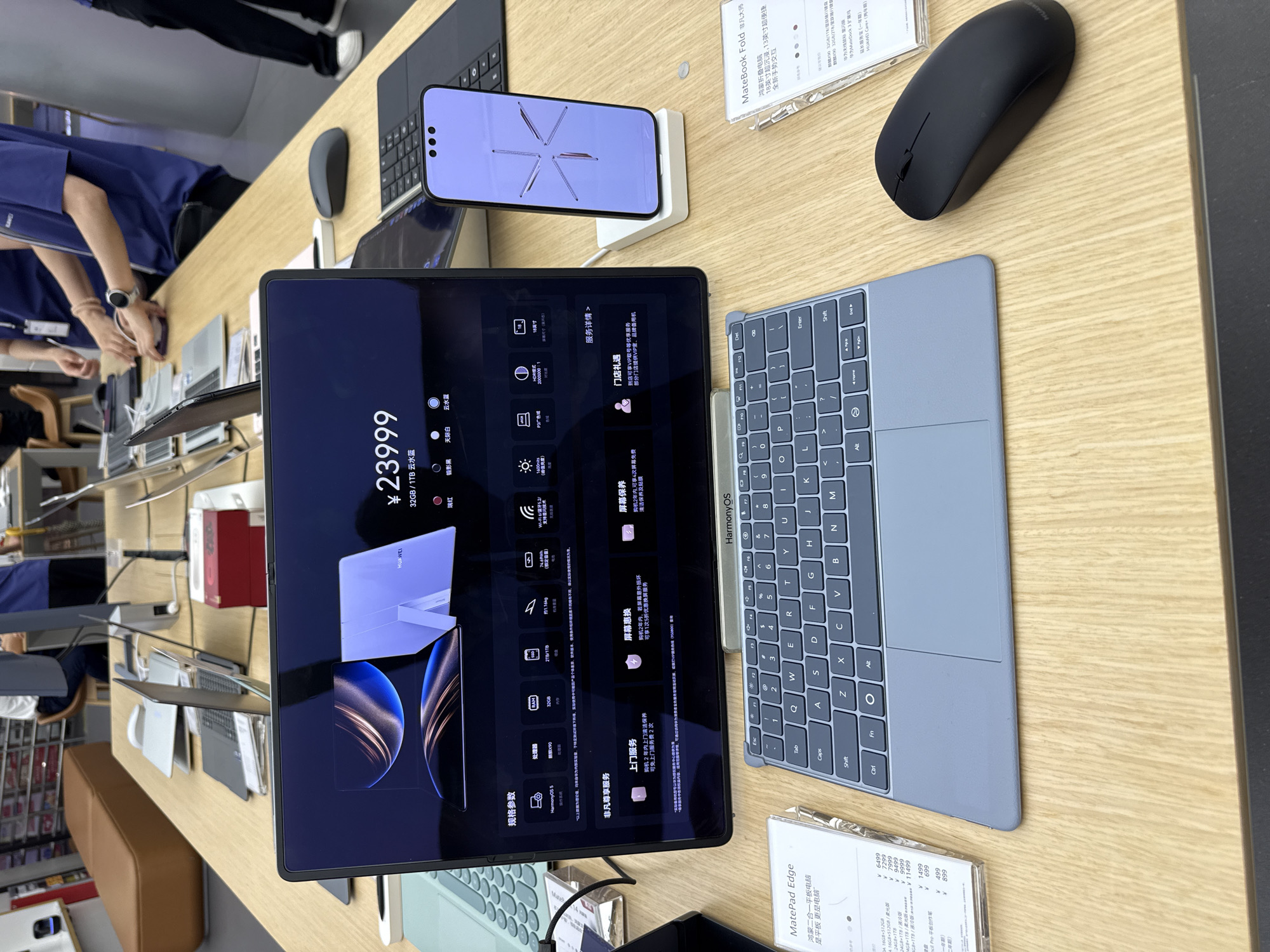 A well-lit retail display showcases Huawei devices. A black Huawei MatePad Pro is centrally positioned, displaying a price of ¥23999 and specifications. A Huawei MatePad keyboard, wireless mouse, and Huawei MateBook Fold stand nearby. A person in a blue shirt appears to be interacting with the display. Several similar devices are displayed in the background. In the background is a retail store, possibly in the Yuexiu District of China.