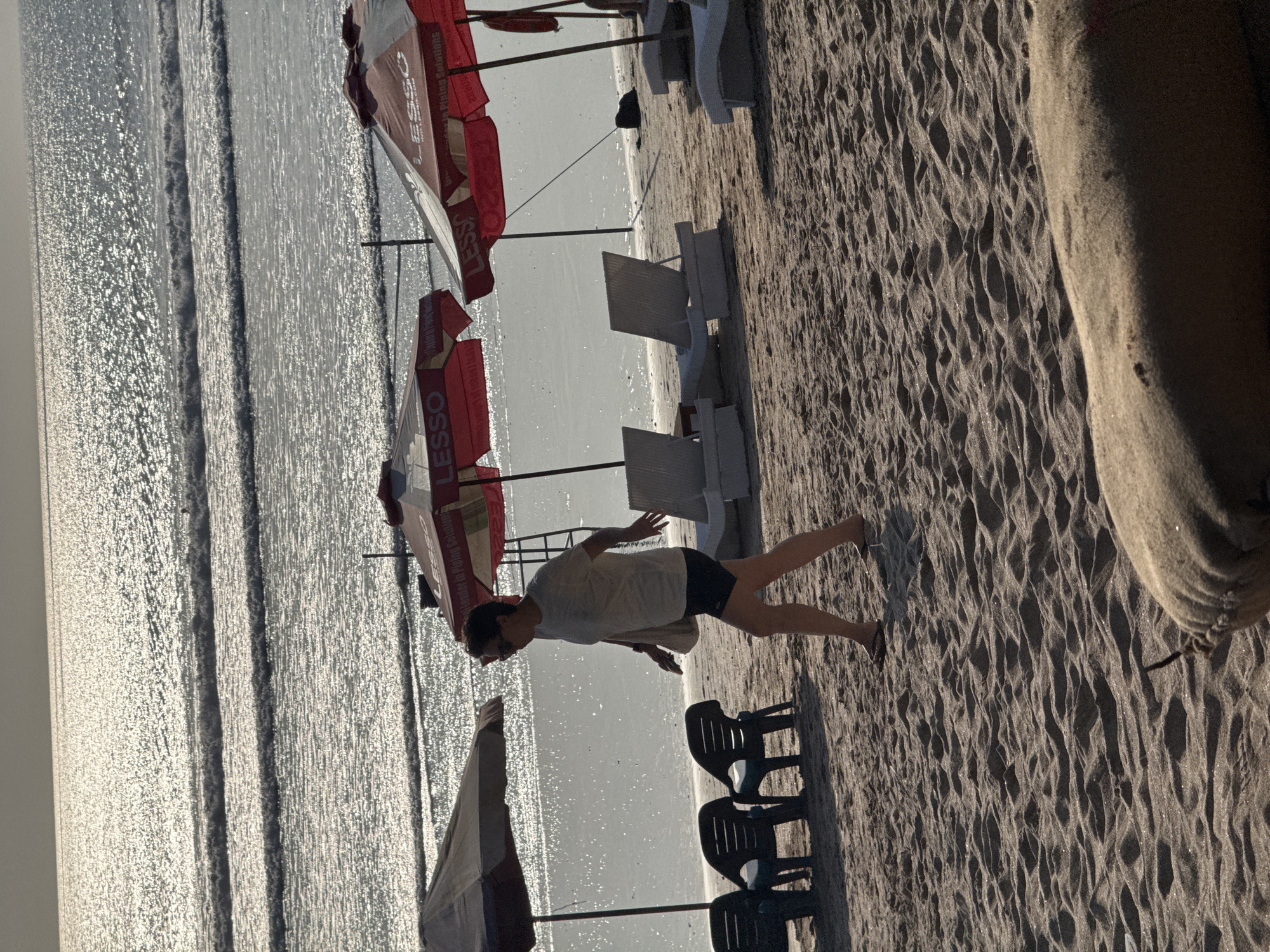 A solitary woman walks along the sandy shore of Kuta Beach in Bali, Indonesia. She is wearing a light-colored t-shirt, dark shorts, sunglasses, and flip-flops. Her hair is pulled back, and she carries a bag over her shoulder. The sun glints off the ocean waves, creating a shimmering effect. In the background, beach chairs and red and white umbrellas are set up on the sand. The sand itself is textured with ripples from the wind and tide. The overall scene is one of a peaceful afternoon at a popular tourist destination.