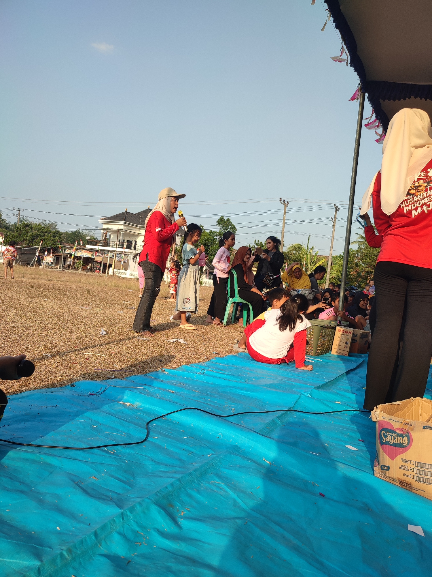 In Nusa Jaya, Indonesia, an outdoor event is taking place under a clear blue sky. A woman wearing a red shirt, headscarf, and cap holds a microphone, appearing to address a gathering. She stands on a field of dry grass, with a large blue tarp spread out in the foreground.
Several children and adults are visible. Some children are sitting on the blue tarp, while others stand around. A woman in a white headscarf and red shirt is partially visible on the right, facing away from the camera. In the background, there are buildings, trees, and utility poles. The overall scene suggests a community gathering or performance. A cardboard box with "Sayang" branding is placed on the blue tarp.