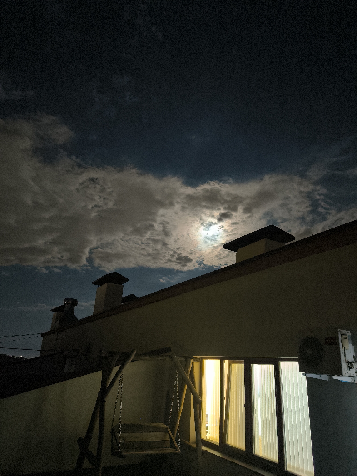 An outdoor nighttime scene depicts a rooftop with a balcony in the city of Chișinău, Moldova. The sky is dark, with scattered clouds illuminated by the moon, which is visible through a break in the clouds. A building's roof, with two chimneys, extends across the lower part of the frame. Below the roof, a wooden swing set is positioned on a balcony. To the right, large windows with vertical blinds are illuminated from within. An outdoor air conditioning unit is mounted on the wall above the windows. The overall impression is of a quiet, residential area at night.