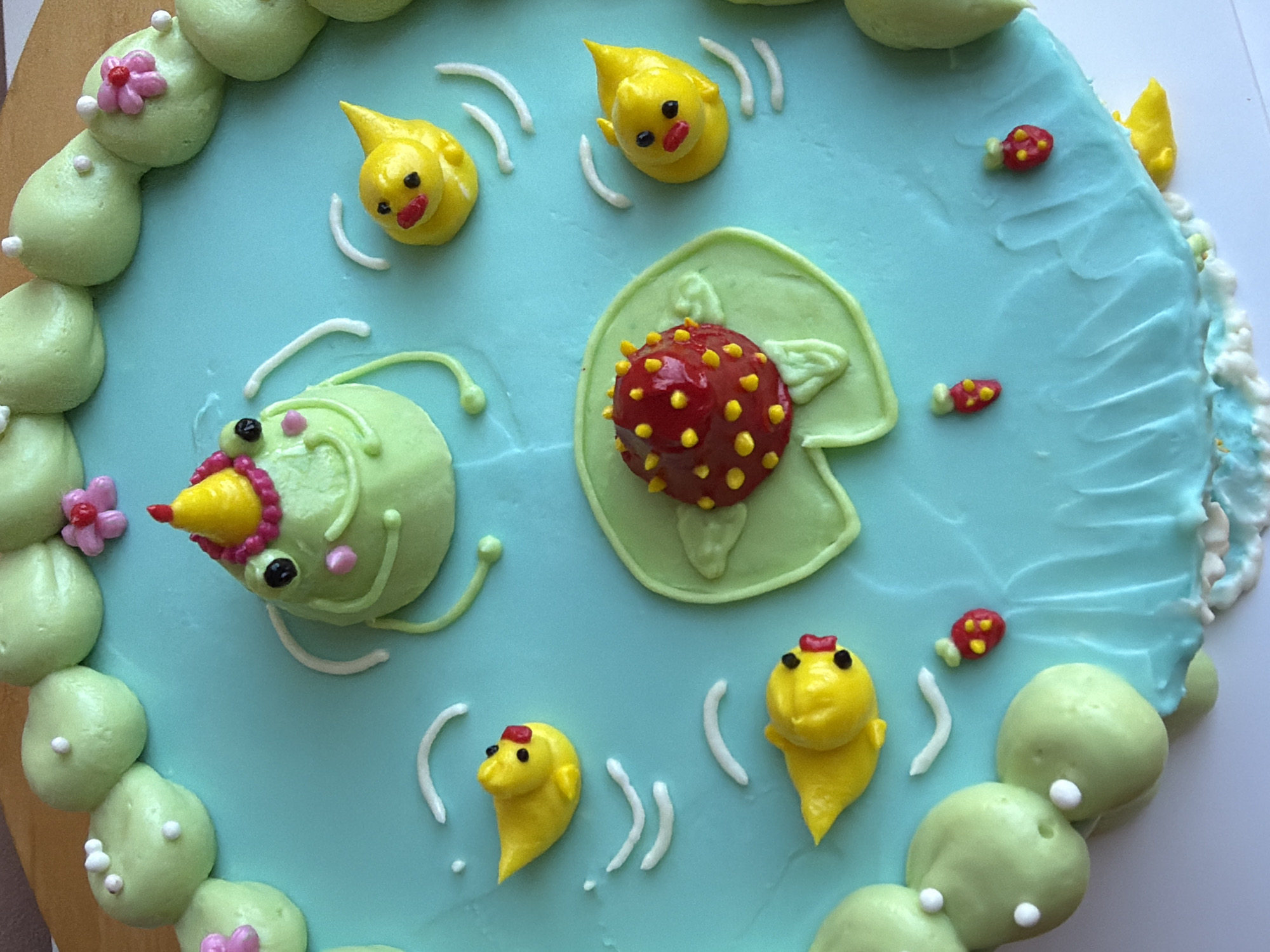 This media file displays a close-up, overhead view of a round cake decorated with a light blue frosting representing water. The cake is adorned with various fondant or buttercream figures, including several yellow ducklings with red accents on their heads, small red fish with yellow spots, and a green frog-like creature wearing a yellow cone hat and sporting pink cheeks and black eyes. There is also a large, spiky red sphere with yellow dots, possibly representing a frog spawn or a ladybug, placed on a green lily pad. The cake's border is decorated with pale green mounds that resemble pebbles or leaves, some with tiny white dots and pink flowers. White icing is used to create wavy lines on the blue surface, suggesting ripples or movement in the water. The overall scene evokes a whimsical, aquatic theme, likely intended for a child's celebration. The lighting suggests indoor illumination, and there are no visible textual elements in the image. The setting is likely a home or a bakery in Tel-Aviv, Israel, given the provided location context.