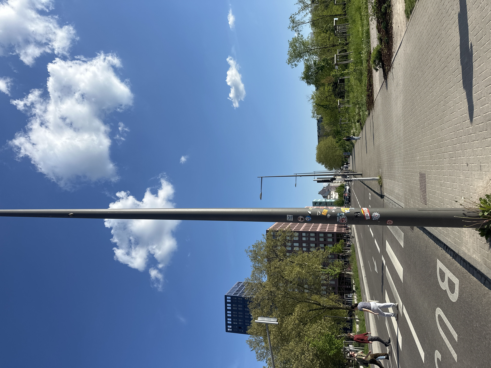 The image depicts a street scene in Strasbourg, France, under a clear, bright blue sky with scattered cumulus clouds. A grey cylindrical pole runs diagonally across the lower half of the frame, adorned with numerous stickers at its upper portion. In the background, there are modern buildings, including a red brick building and a dark blue high-rise, interspersed with green trees. The foreground shows a crosswalk with the word "BUS" painted on the asphalt. Three people are walking across the crosswalk. A paved walkway runs along the right side of the street. Further in the background, a line of evenly spaced streetlights extends down the street.