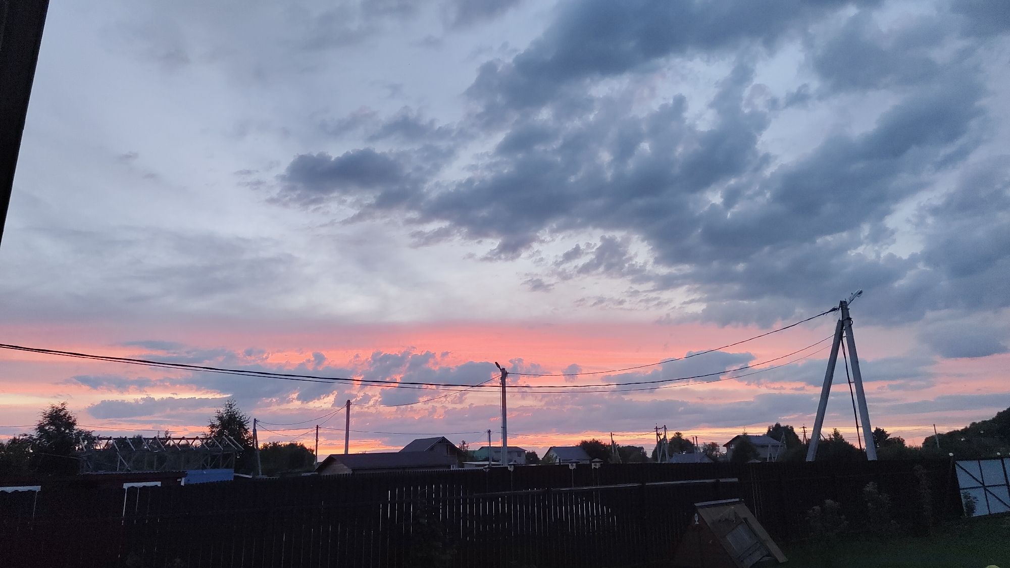 The image depicts a rural scene at dusk in Rybolovo, Russia. The sky is a dramatic canvas of clouds, with a band of vibrant pink and orange hues from the setting sun illuminating the horizon. Above this, the clouds are a mix of grey and lighter shades, creating depth and texture. Several utility poles with power lines crisscross the view, silhouetted against the colorful sky. In the foreground, a dark wooden fence runs horizontally across the frame. Behind the fence, the roofs of several houses and a structure under construction are visible, partially obscured by trees and foliage. The overall impression is one of peaceful, twilight stillness.