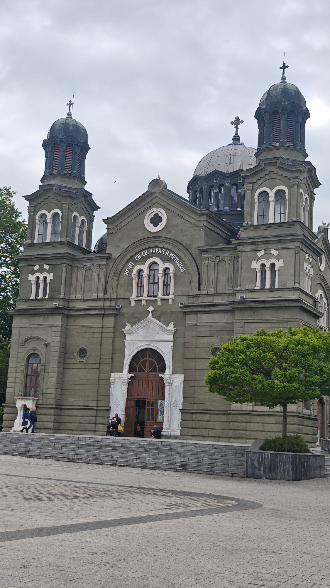 The image depicts the Church of Saints Cyril and Methodius in Burgas, Bulgaria. The church is built of light gray stone with two prominent towers and a central dome. The text "ХРАМ.. СВ. СВ. КИРИЛ И МЕТОДИЯ" is inscribed above the main entrance. A few people are visible on the steps leading to the church entrance. The scene is set on a cloudy day with trees and a large paved area in front of the church.