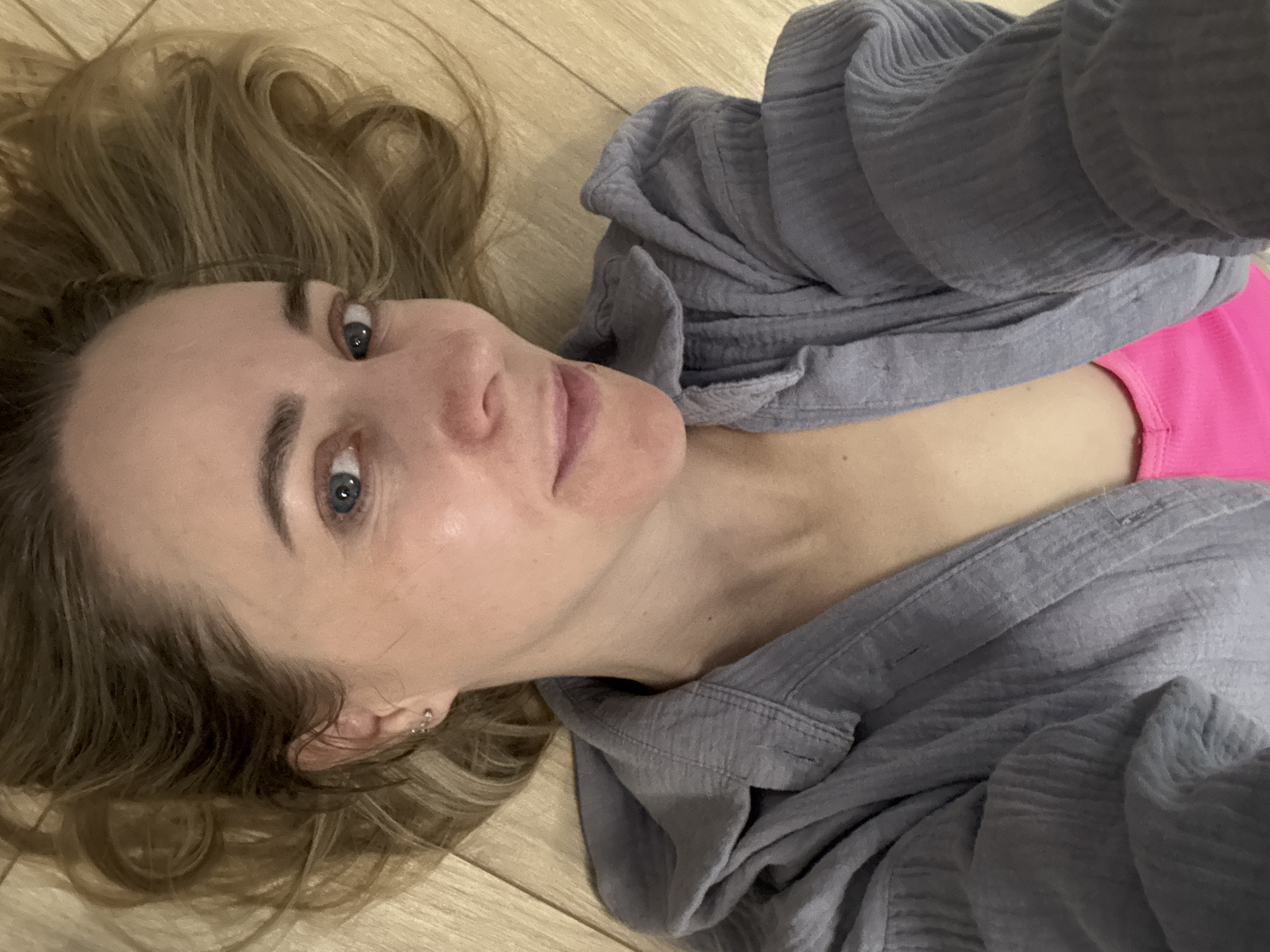 A fair-skinned woman with long, wavy blonde hair is lying on a light wood floor. She is wearing a gray button-down shirt over a bright pink top. The location is unknown.