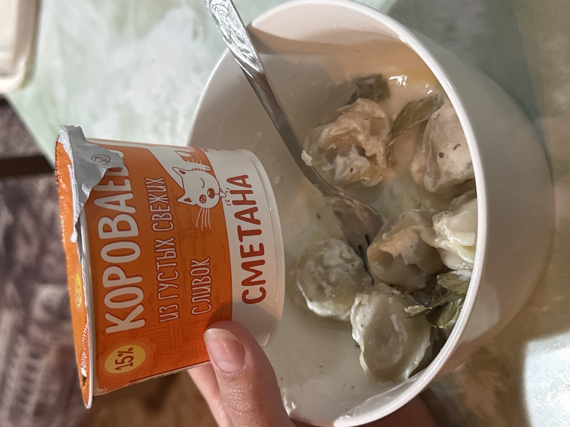 In this close-up shot, a person's hand is holding an open container of "Короваево" brand sour cream, with 15% fat content indicated. The sour cream is being poured into a white bowl filled with dumplings in a creamy sauce, with some green herbs also present. A spoon is in the bowl, ready to mix or serve the food. The background is slightly out of focus, showing a table with a textured surface and hints of a light green and white patterned tablecloth. The lighting suggests it is daytime, possibly indoors. The visible text on the sour cream container is in Russian: "КОРОВАЕВО", "СМЕТАНА", "ИЗ ГУСТЫХ СВЕЖИХ СЛИВОК", and "15%". The scene depicts the preparation or serving of a meal, likely at home. The image is likely taken in Шолохово, Russia, based on the location context.