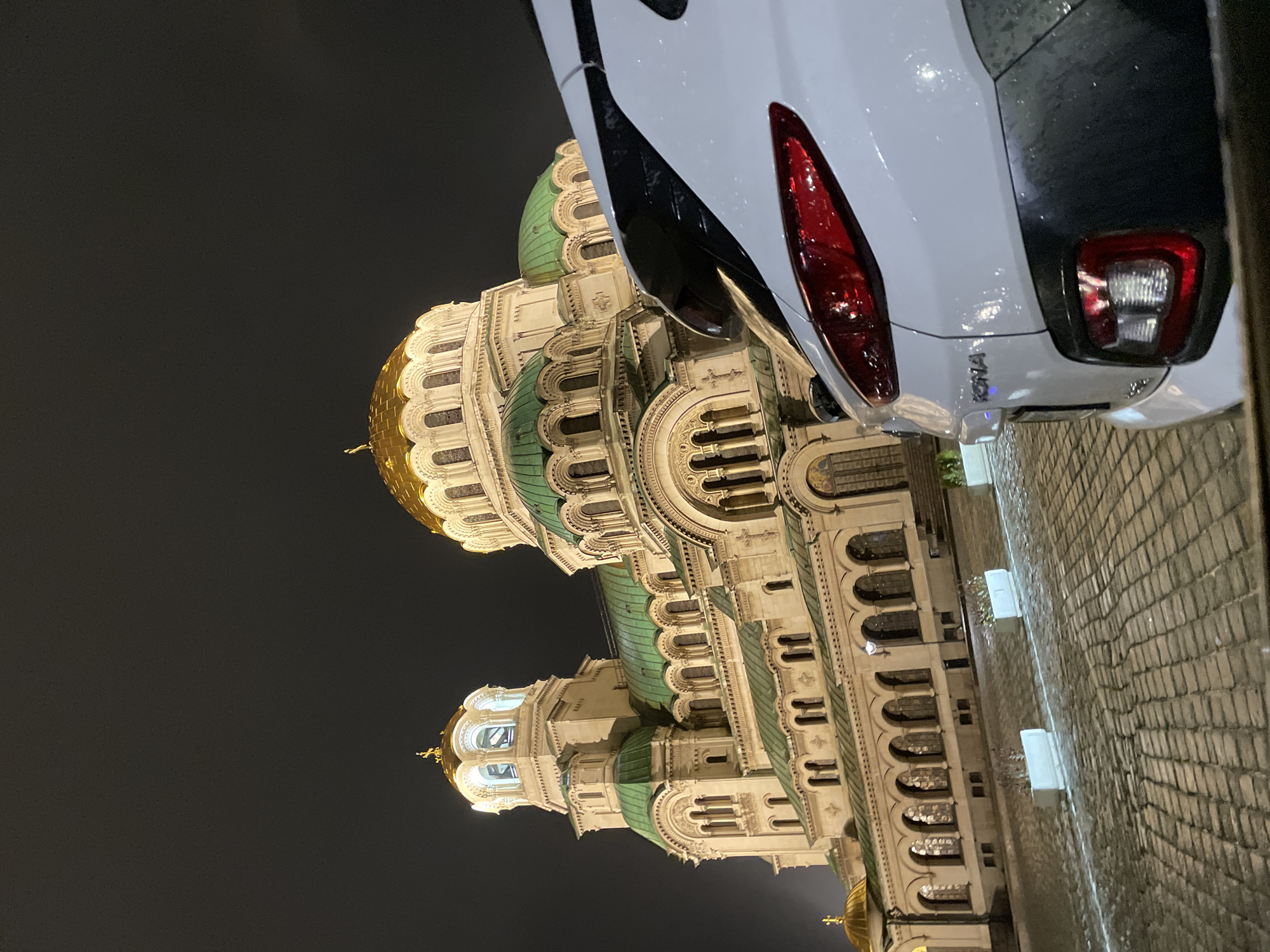 The image shows the St. Alexander Nevsky Cathedral in Sofia, Bulgaria, at night. The cathedral's ornate architecture, featuring green domes and golden accents, is illuminated against the dark sky. A white car, a Hyundai Kona, is parked partially in front of the cathedral, with its rear end visible in the foreground. The ground is paved with cobblestones and appears wet, suggesting it has recently rained. No people are visible in the frame.