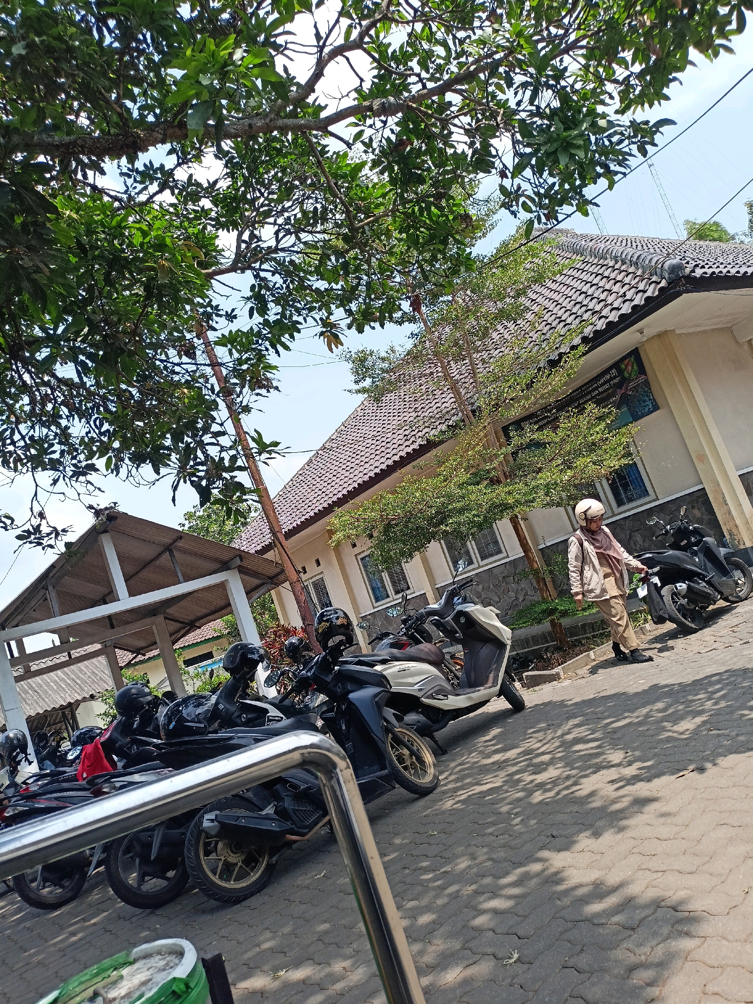 In Kewaluhan, Indonesia, a street scene unfolds under a clear blue sky, with the dappled shade of trees falling across a paved walkway. Several parked motorcycles, predominantly black and white, are lined up, some with helmets resting on their seats. To the right, a woman wearing a tan hijab and matching tan clothing walks near a light-colored building with a tiled roof. The building features multiple windows and a sign with text. In the background, another covered structure with a corrugated metal roof is visible. The overall impression is one of a tranquil, everyday setting in an Indonesian town.