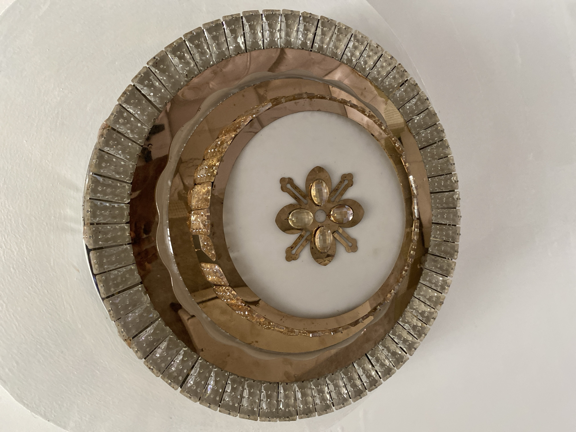 This image shows a close-up, top-down view of an ornate, circular decorative piece mounted on a white ceiling. The object appears to be a ceiling fixture or embellishment, possibly a lamp or a purely decorative element.
The object itself is layered and consists of several concentric rings. The outermost ring is a band of clear, faceted crystals, arranged vertically and closely packed, creating a glittering edge. Inside this, there are bands of what appear to be mirrored or metallic surfaces with a warm, coppery or brassy hue. These inner rings have a curved or wavy texture in places.
At the very center of the object is a small, metallic embellishment shaped like a stylized flower or star. This central piece features what look like clear or light amber colored stones set within its design.
The overall impression is one of opulence and intricate craftsmanship. The lighting suggests it could be daytime, as the white ceiling is evenly lit, though the source of light is not visible. The setting is indoors, on a ceiling, likely in a residential or public space that values decorative architecture. No people or activities are visible. There is no visible text in the image.