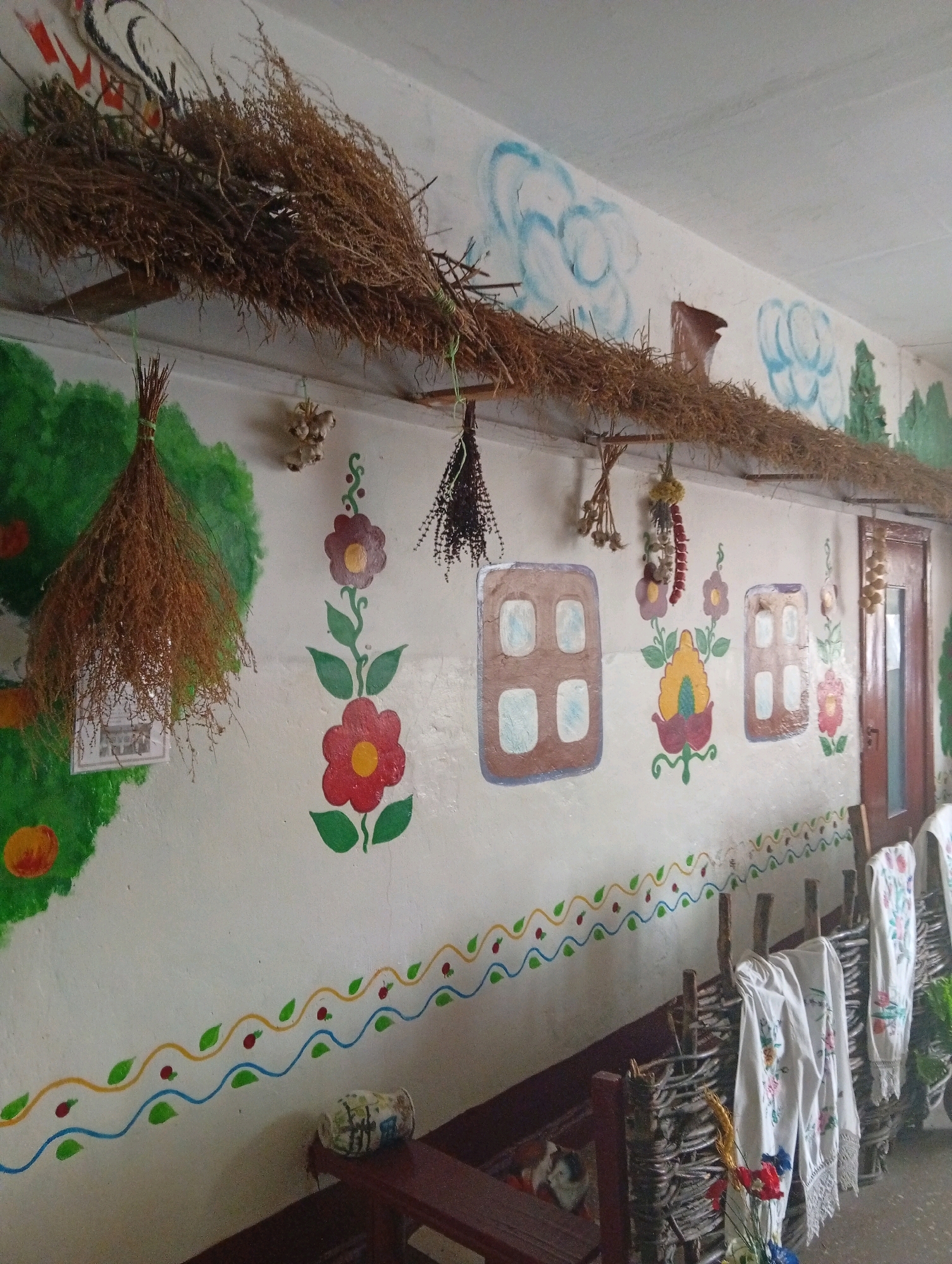 The image depicts the interior of a room decorated in a folk art style, likely a cultural exhibit or a themed space. The walls are painted white and adorned with colorful murals of flowers, trees, and what appear to be stylized clouds. A thatched roof-like structure extends horizontally along the upper portion of the wall, supported by wooden beams. Various dried herbs and plants are hung from the ceiling and the wall, including bunches of dried grass, garlic, and peppers.
Several painted elements mimic architectural features, such as two windows with brown frames and light blue panes. A decorative border of yellow waves, green leaves, and red berries runs along the lower part of the wall.
In the foreground, a rustic woven fence made of branches is visible. White embroidered towels with floral patterns are draped over the fence. To the left, a wooden bench is positioned against the wall. On the bench rests a decorative cylindrical object, possibly a vase, and a small section of a rug or fabric with what might be a cat's tail.
The lighting appears to be from artificial sources, casting a consistent illumination across the scene. The overall impression is one of a handcrafted, traditional ambiance.