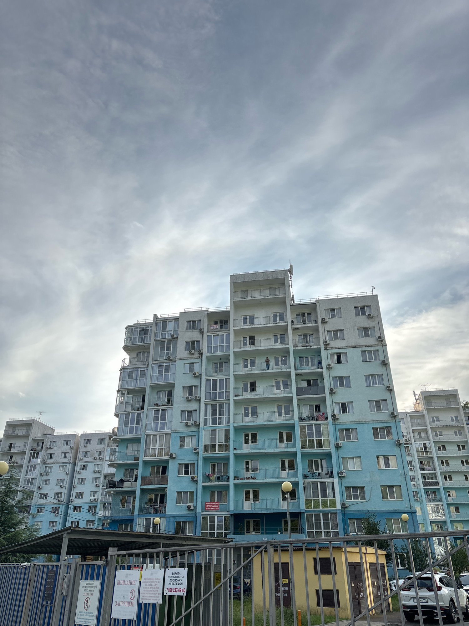 The image shows a large, light blue apartment building with multiple stories and many windows, located in the city of Razdolnoye, Russia. The building has balconies on various floors and air conditioning units are visible outside many windows. The sky above is overcast with grey clouds. In the foreground, there is a metal fence with several signs attached to it. Behind the fence, there is a small yellow structure with dark doors, and a white SUV is parked to the right. Other apartment buildings, partially visible, flank the main building. The overall scene depicts a residential area under a cloudy sky.